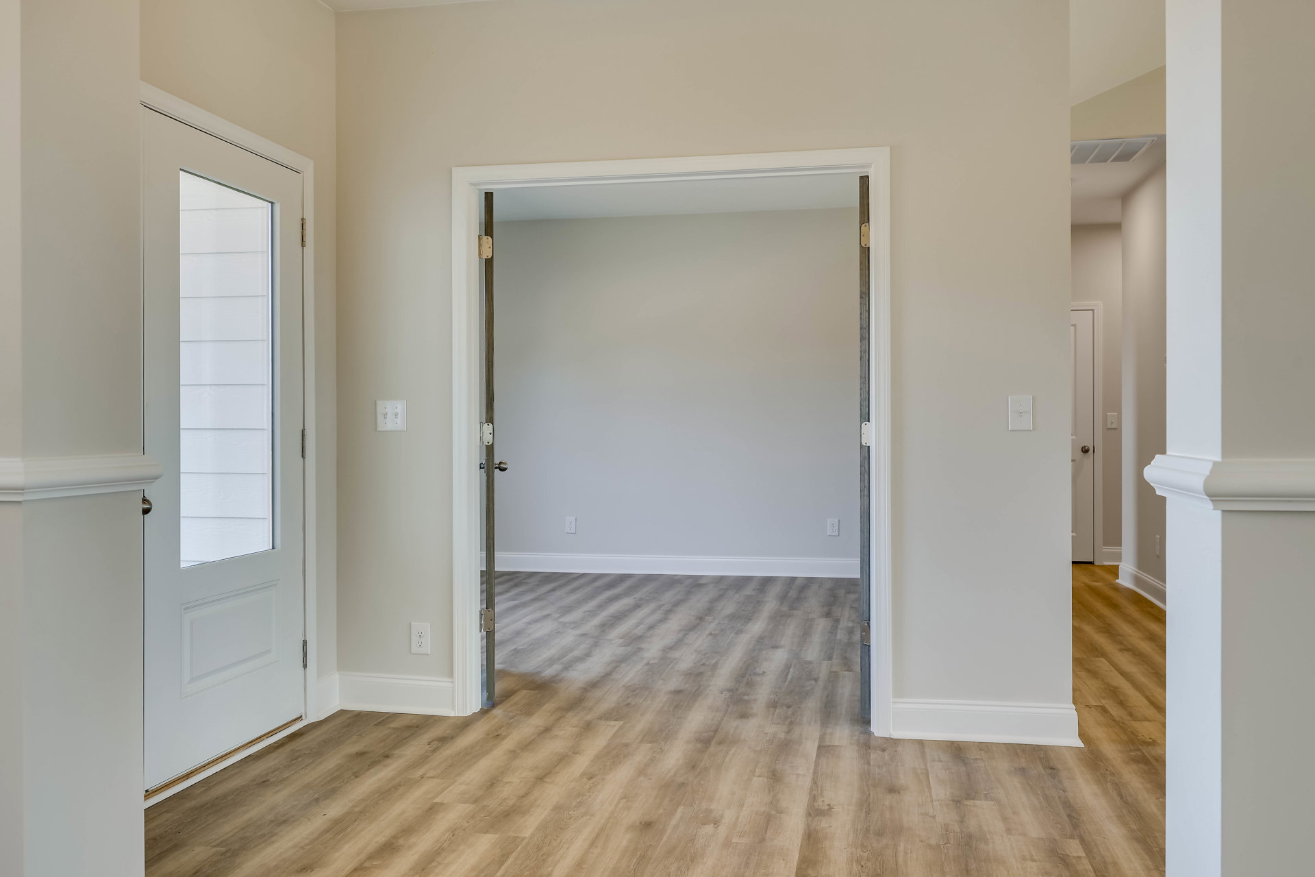 White paneled door open to a room with light wood flooring, white walls, and a visible corner; refrigerator and cabinetry partially seen in the background.