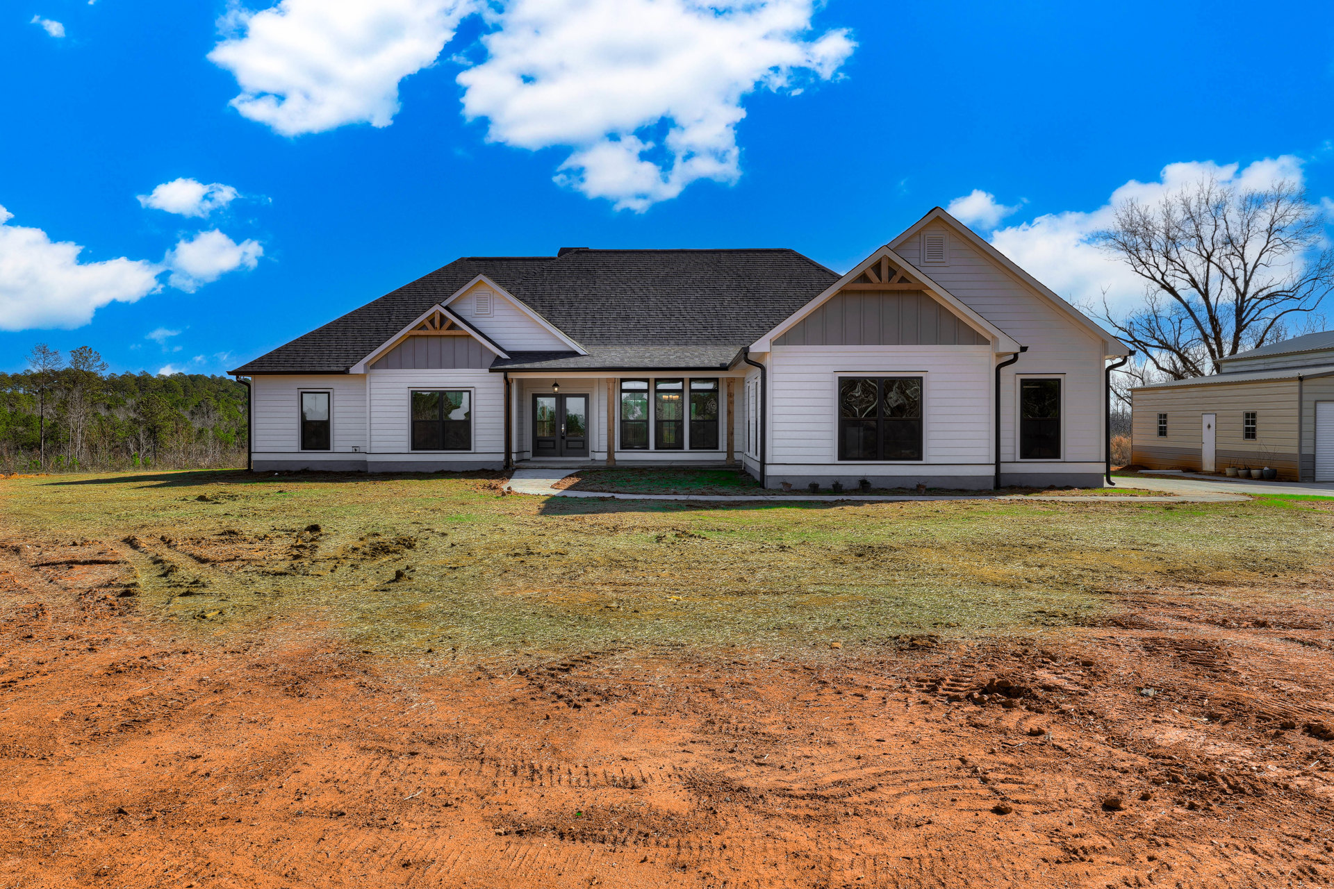 White house with black roof and black-framed windows, surrounded by green lawn and small trees under a blue sky with scattered clouds