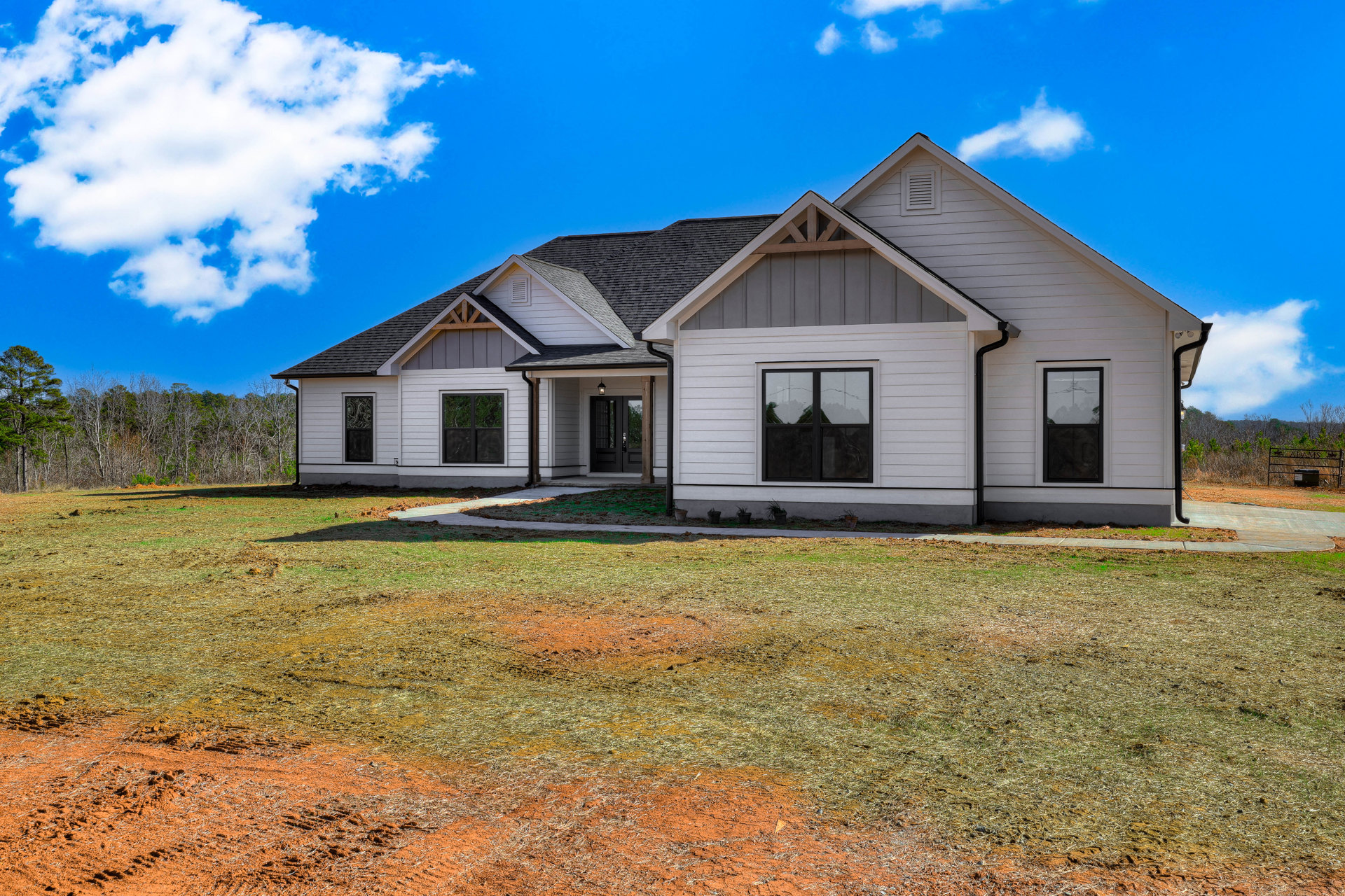 Modern two-story home with black-framed windows, light siding, green lawn bordered by dirt, and blue sky with scattered clouds; neighboring houses visible in the background.