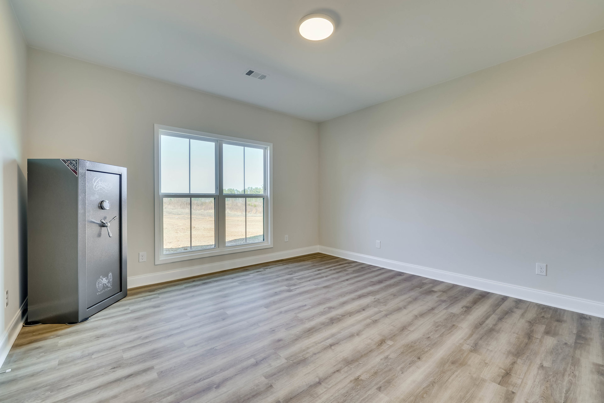 Wood-floored room with a grey combination safe, plaster walls, ceiling light, and window overlooking grassy field