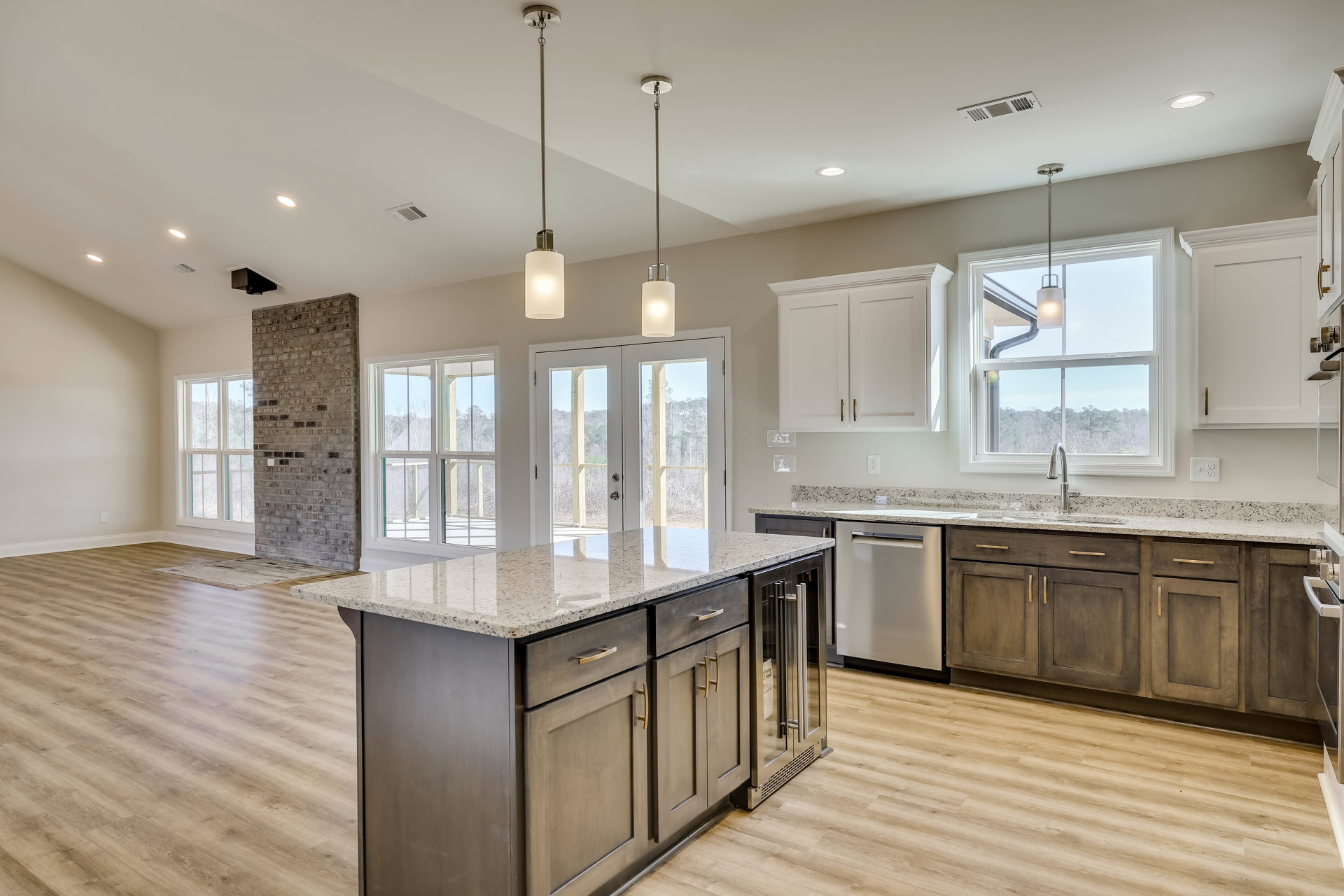 Spacious kitchen featuring a large granite island, wood flooring, white cabinetry, stainless steel sink, brick accent wall with window, and double glass doors