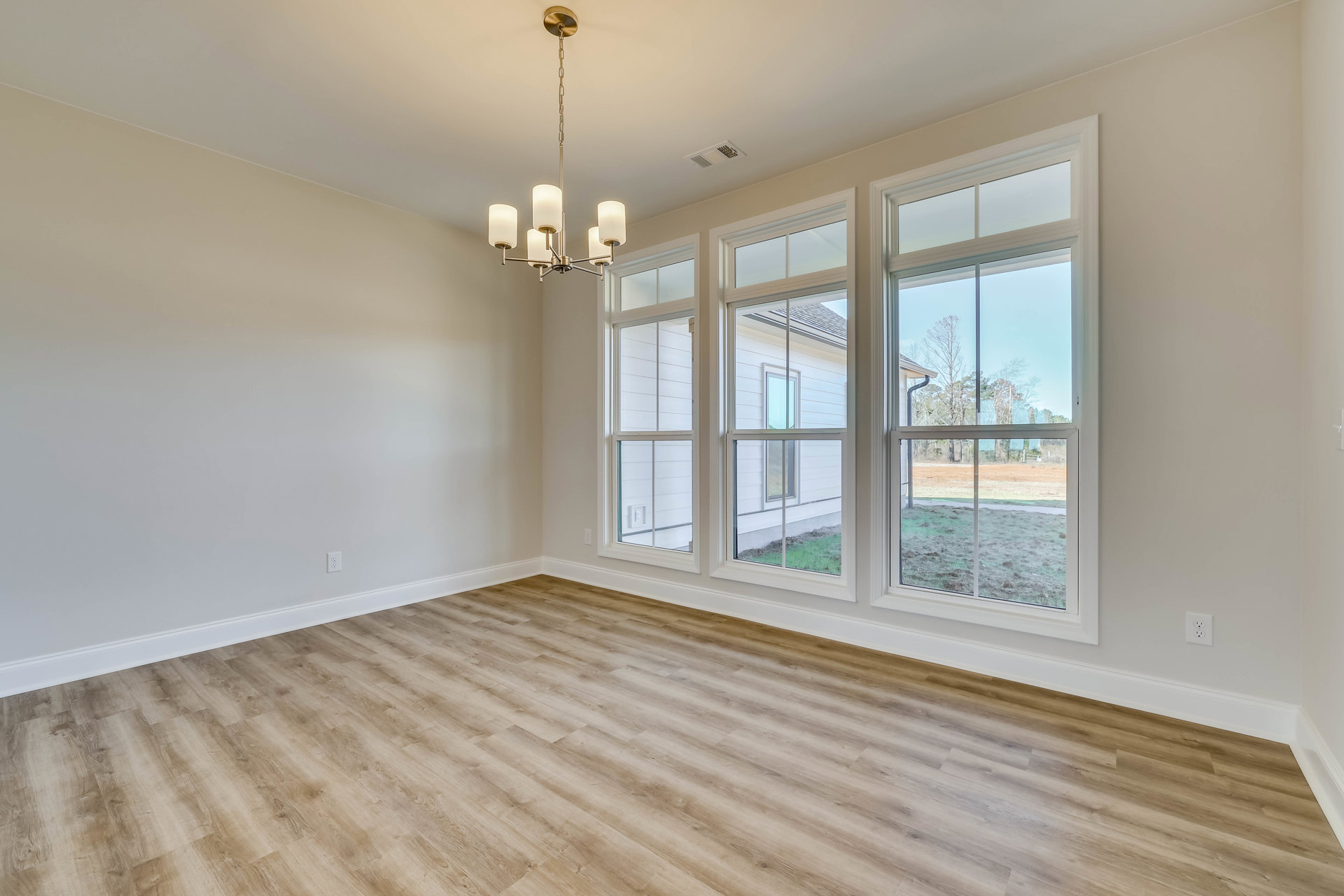 Chandelier hanging from white ceiling above wood flooring, large windows with white frames, view of neighboring house and trees outside