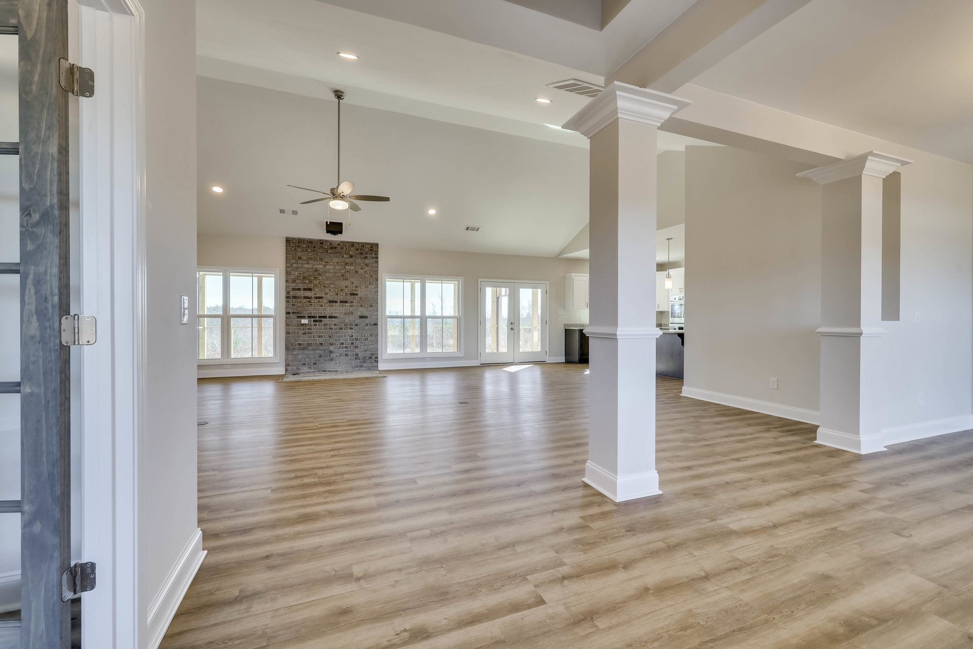 Spacious living area featuring a stone fireplace, wood flooring, white pillars, ceiling fan, and large windows with views of trees outside.