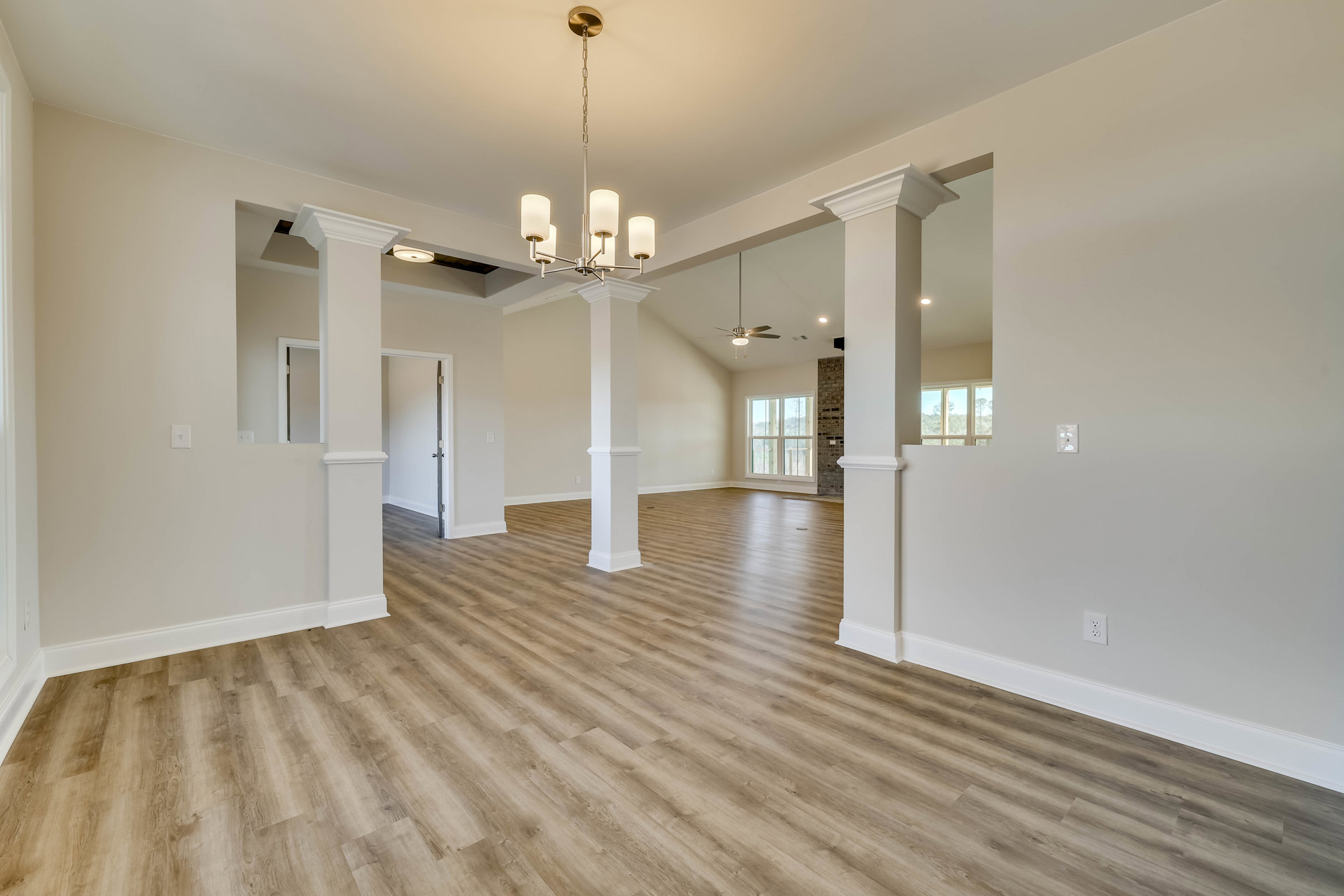 Open room with rich wood flooring, white pillars, elegant chandelier, large window overlooking trees, and white door with silver handle