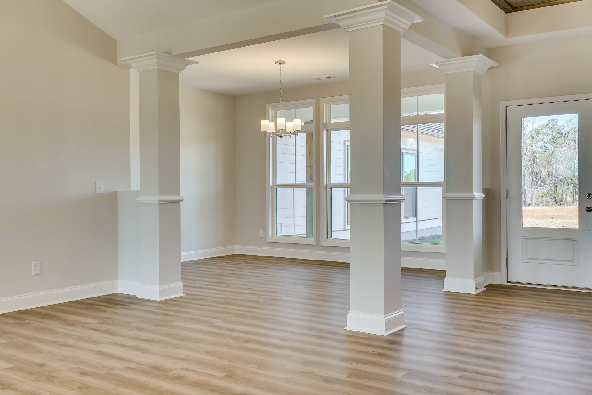 Wood flooring and white columns in a spacious room, white walls, large window with view of trees, glass-paneled door, decorative molding.