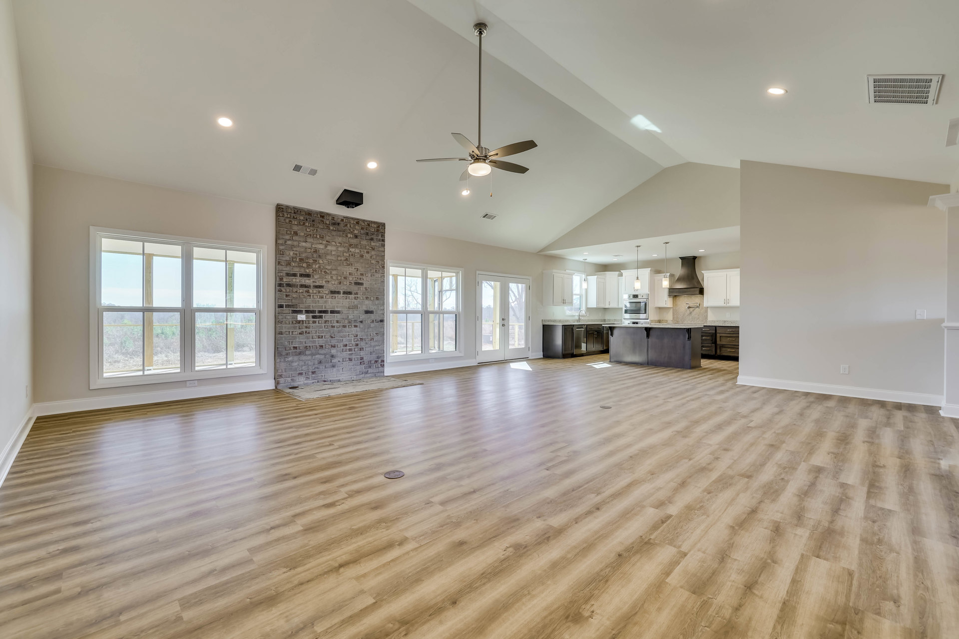 Spacious living area with hardwood floors, brick accent wall featuring a fireplace, ceiling fan, large window offering outdoor views, and visible vent hood and light switch.