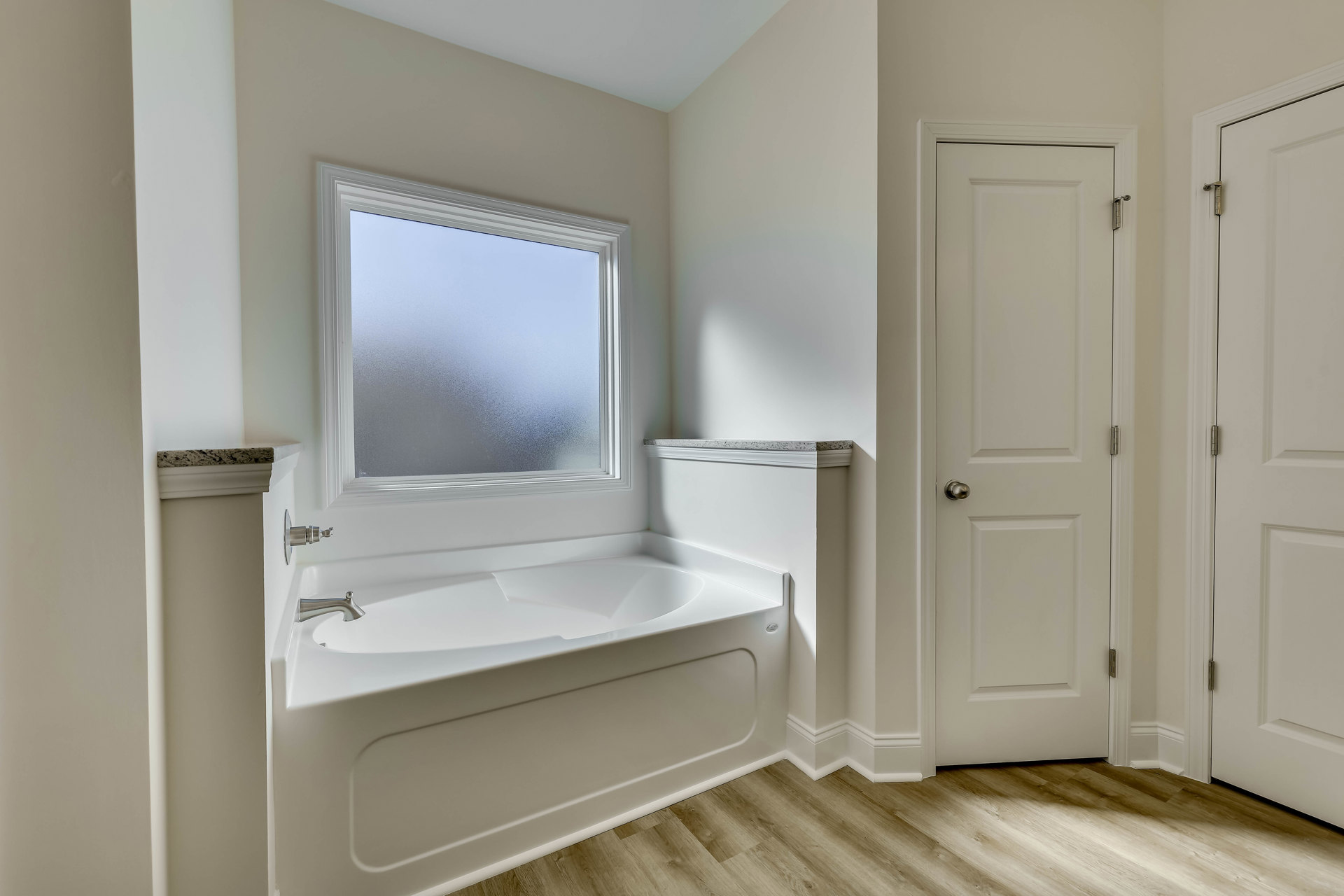 Freestanding white bathtub with smooth surface beneath frosted window, white walls, and silver doorknob on adjacent door