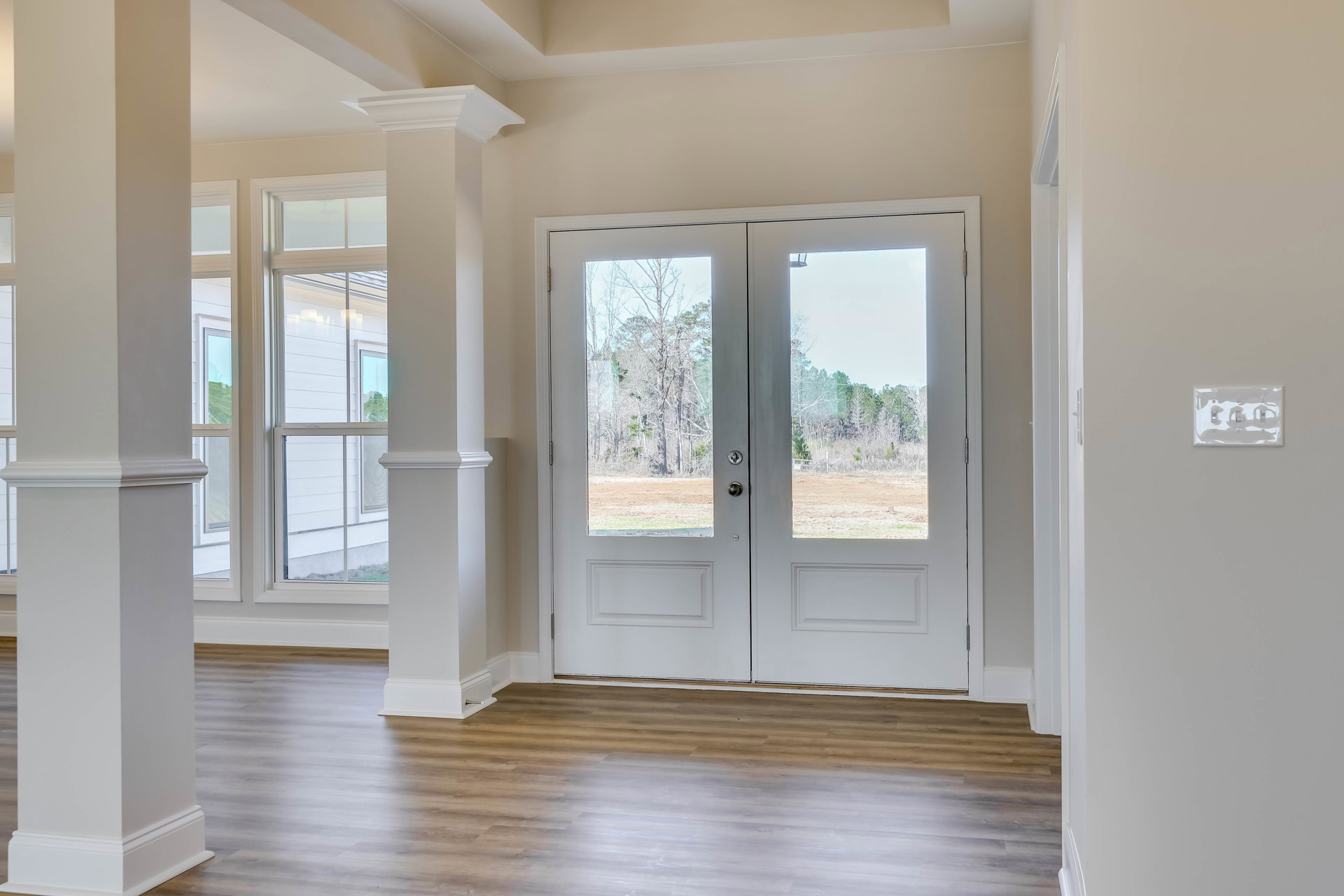 White double doors with glass panes set in a white wall, wood flooring, white-framed window, and light switch visible on the wall.