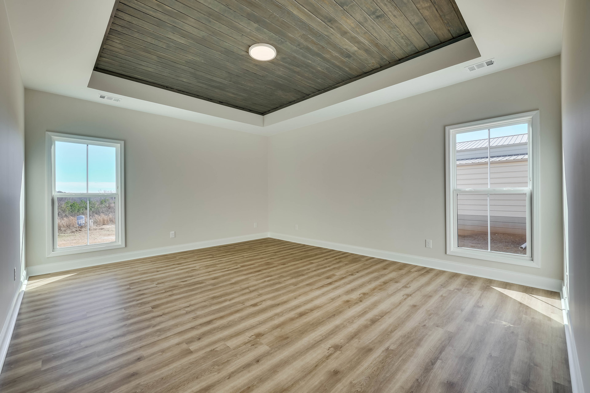Open room with wood plank floor and matching wood ceiling, white walls, large window framed in white overlooking field and trees, modern white light fixture mounted on wood