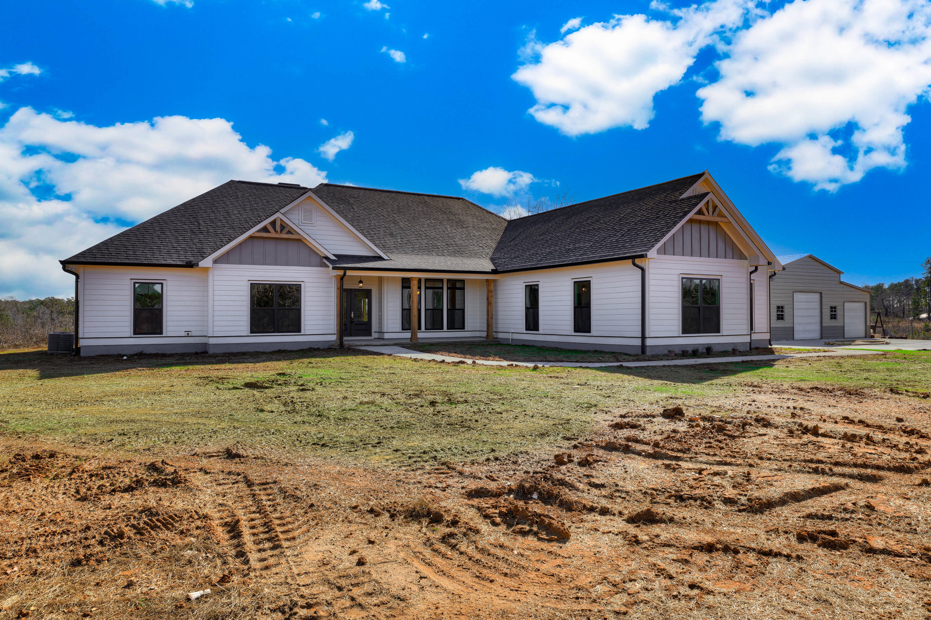 Two-story house with gray siding, covered porch, white trim, large front window reflecting trees, green lawn, and dirt patch under blue sky with scattered clouds