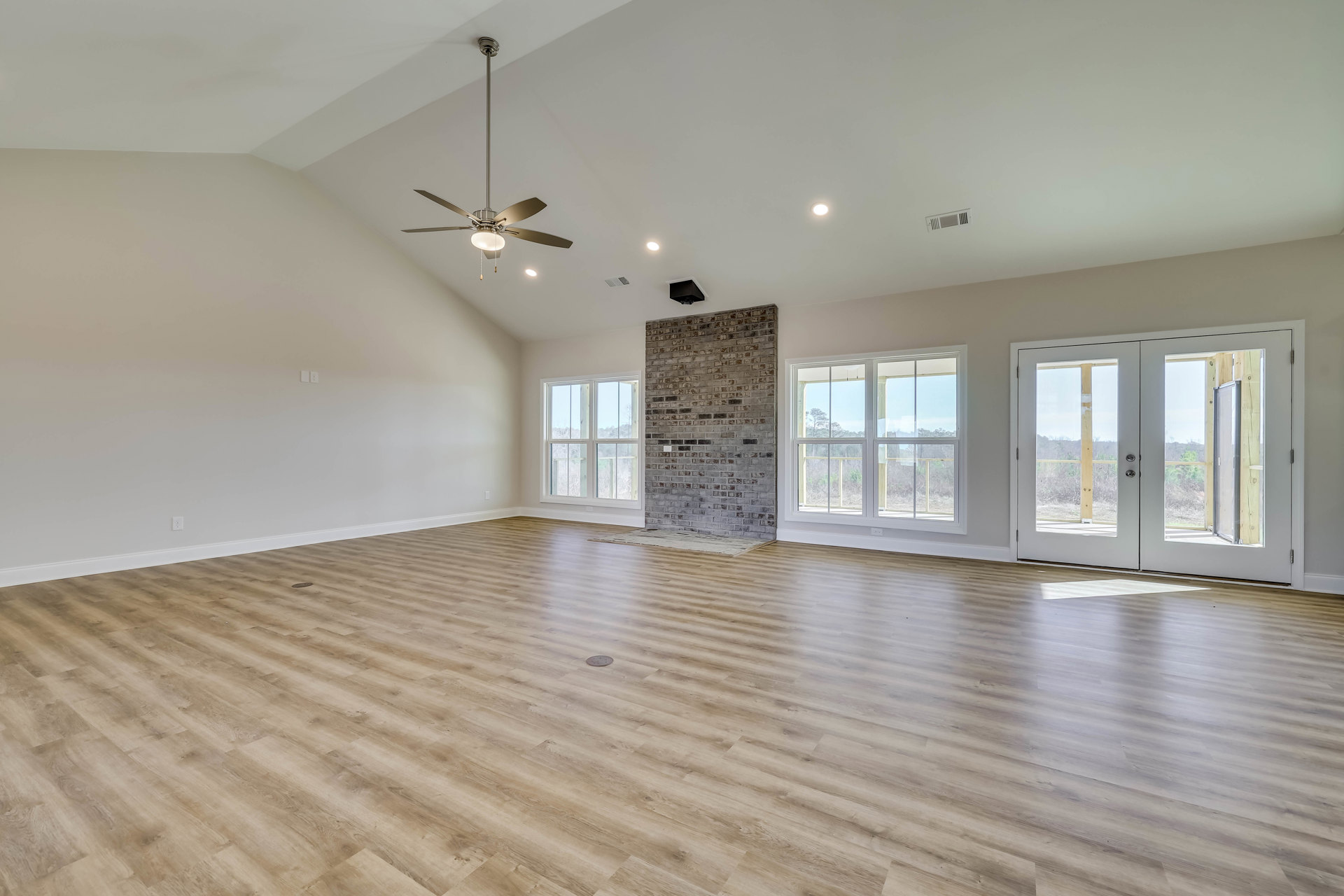 Spacious living area with wood flooring, ceiling fan with light, brick accent wall, white door, fireplace, and large windows overlooking trees and sky