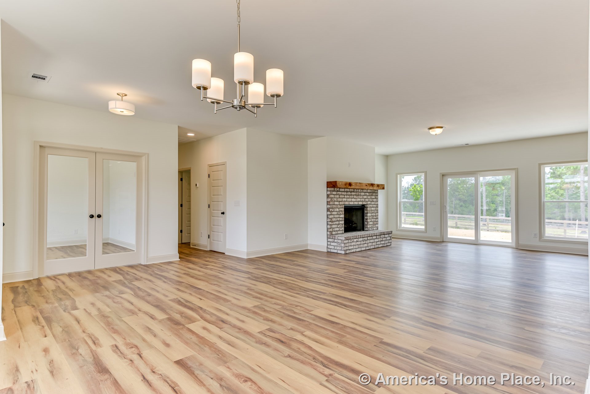 White painted brick fireplace centered in living area with wide plank wood-look flooring, large windows trimmed in white, double glass doors with black handles, modern ceiling