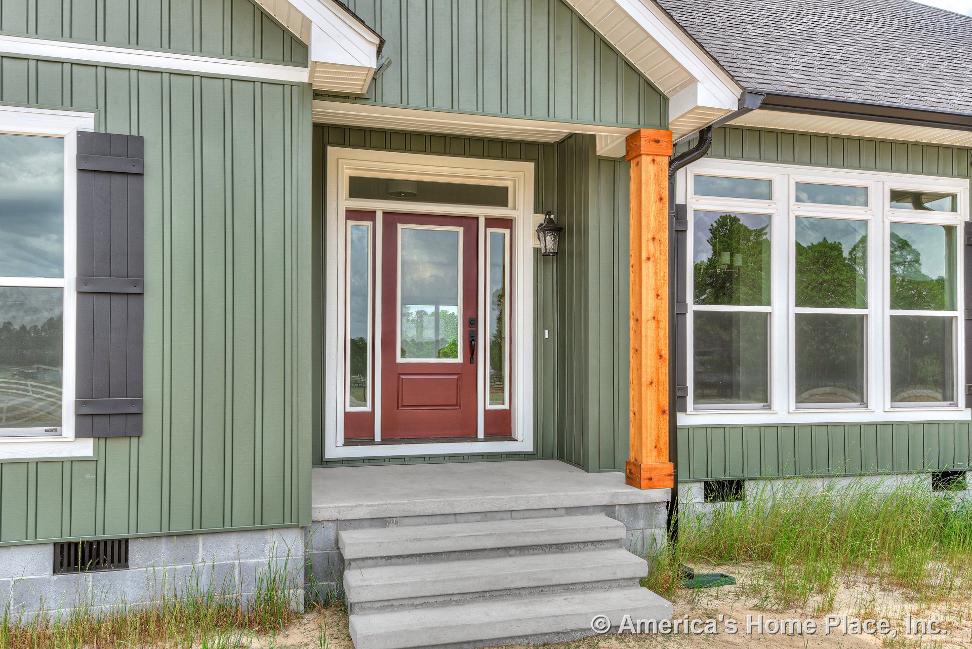 Red front door with glass sidelights, vertical green siding, covered porch with wood support column, concrete steps, white trim windows with shutters, and outdoor lighting