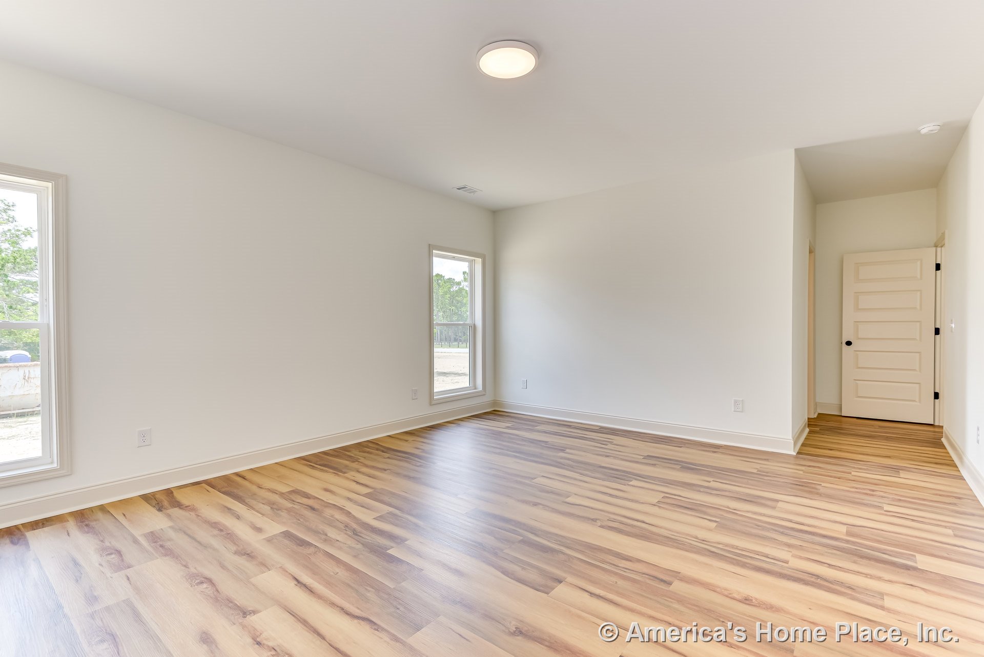Bedroom with light wood-look laminate flooring, white baseboards and trim, two large windows, neutral painted walls, flush mount ceiling light, and paneled interior door in a