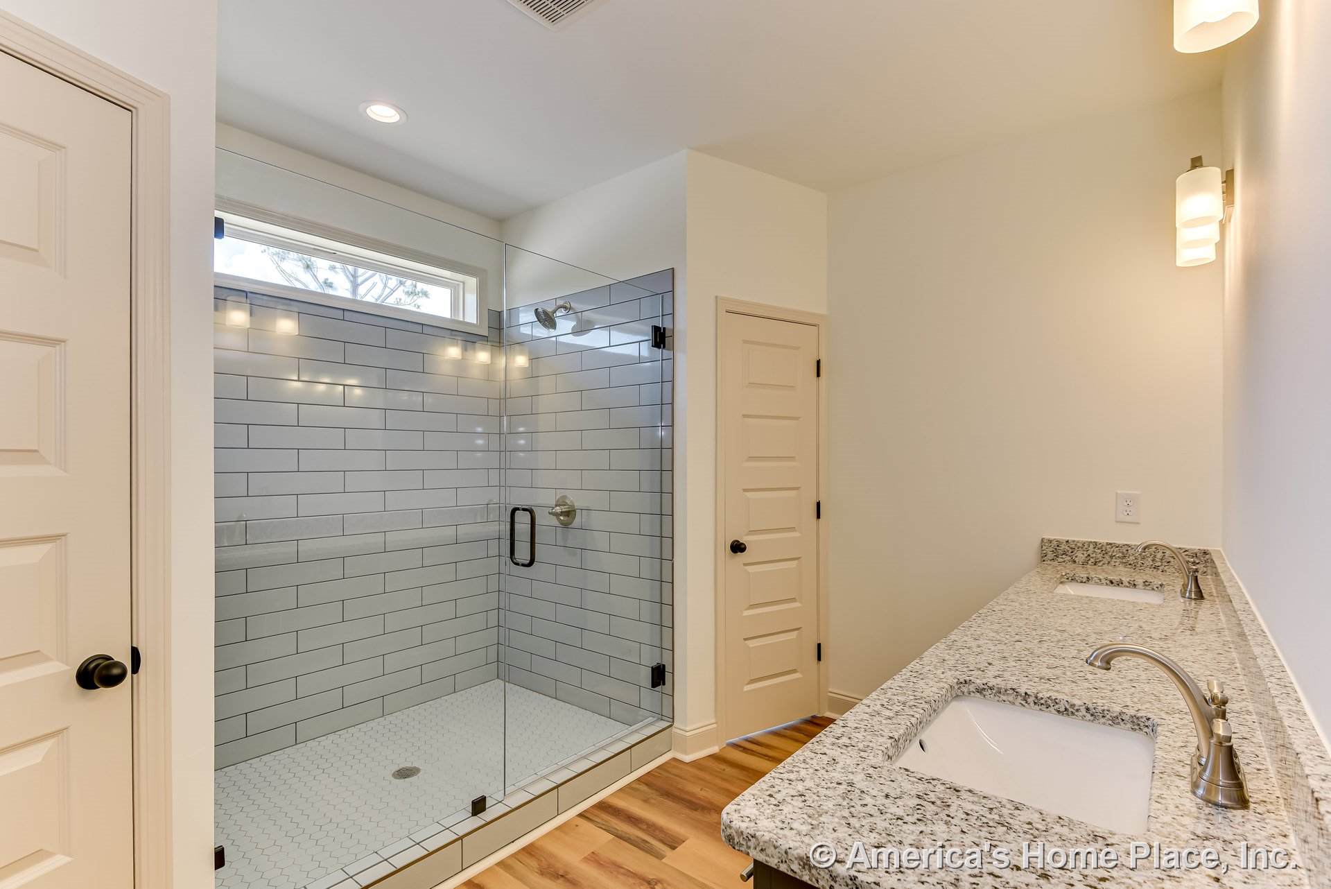 Glass-enclosed shower with white subway tile walls, double vanity featuring granite countertops and undermount sinks, wall-mounted sconces, wood-look flooring, interior door, and