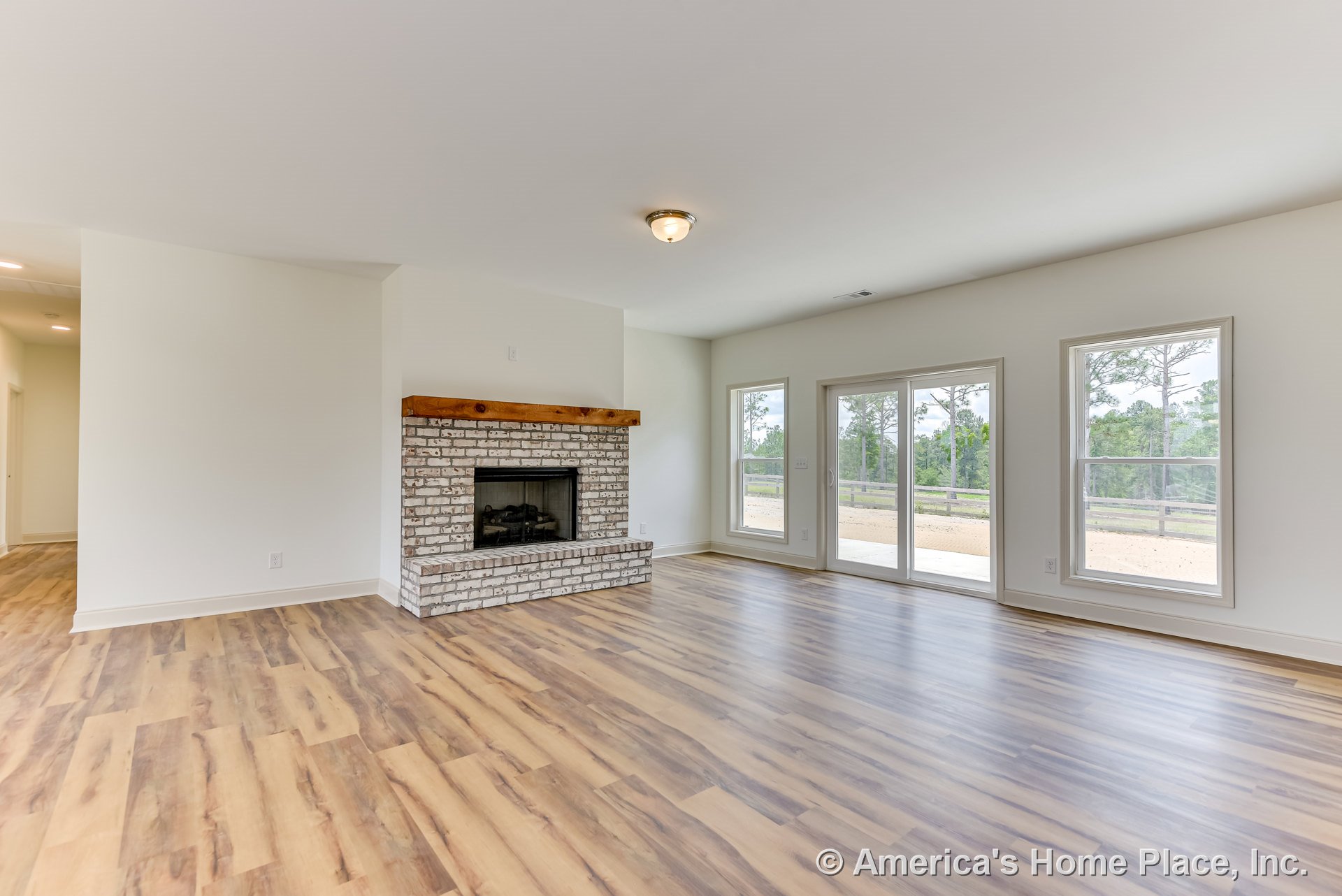 Whitewashed brick fireplace with wood mantel, large windows and glass patio door providing backyard views, light wood flooring, flush ceiling light, and open layout with natural