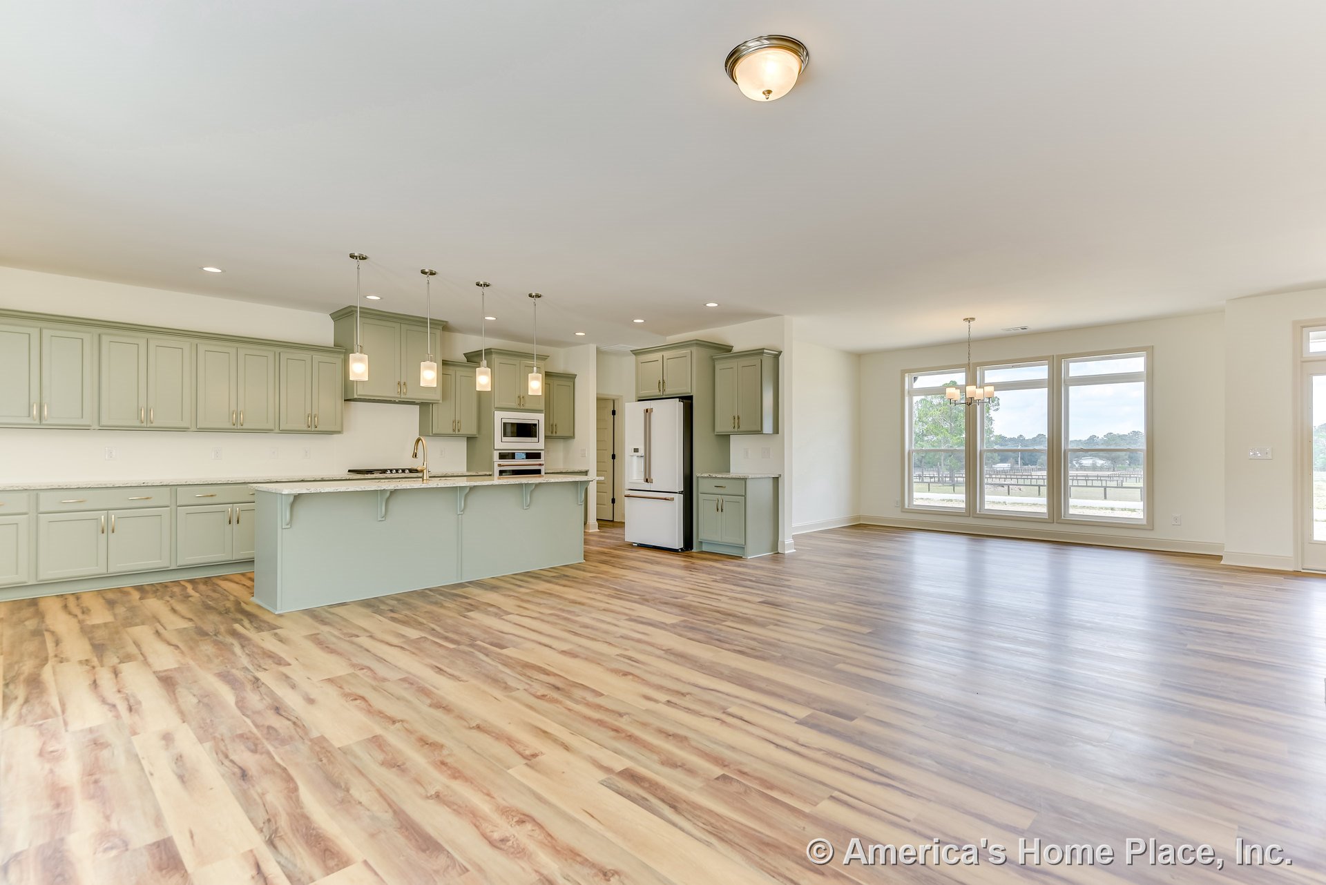 Open concept kitchen and living area with light green cabinetry, large marble island, multiple pendant lights, wide plank wood flooring, expansive windows, built-in oven