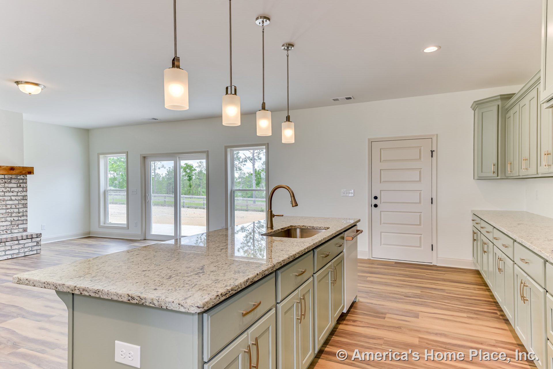 Granite island with undermount sink and multiple pendant lights, painted shaker cabinets with gold hardware, wood-look flooring, brick fireplace with wood mantel, large windows