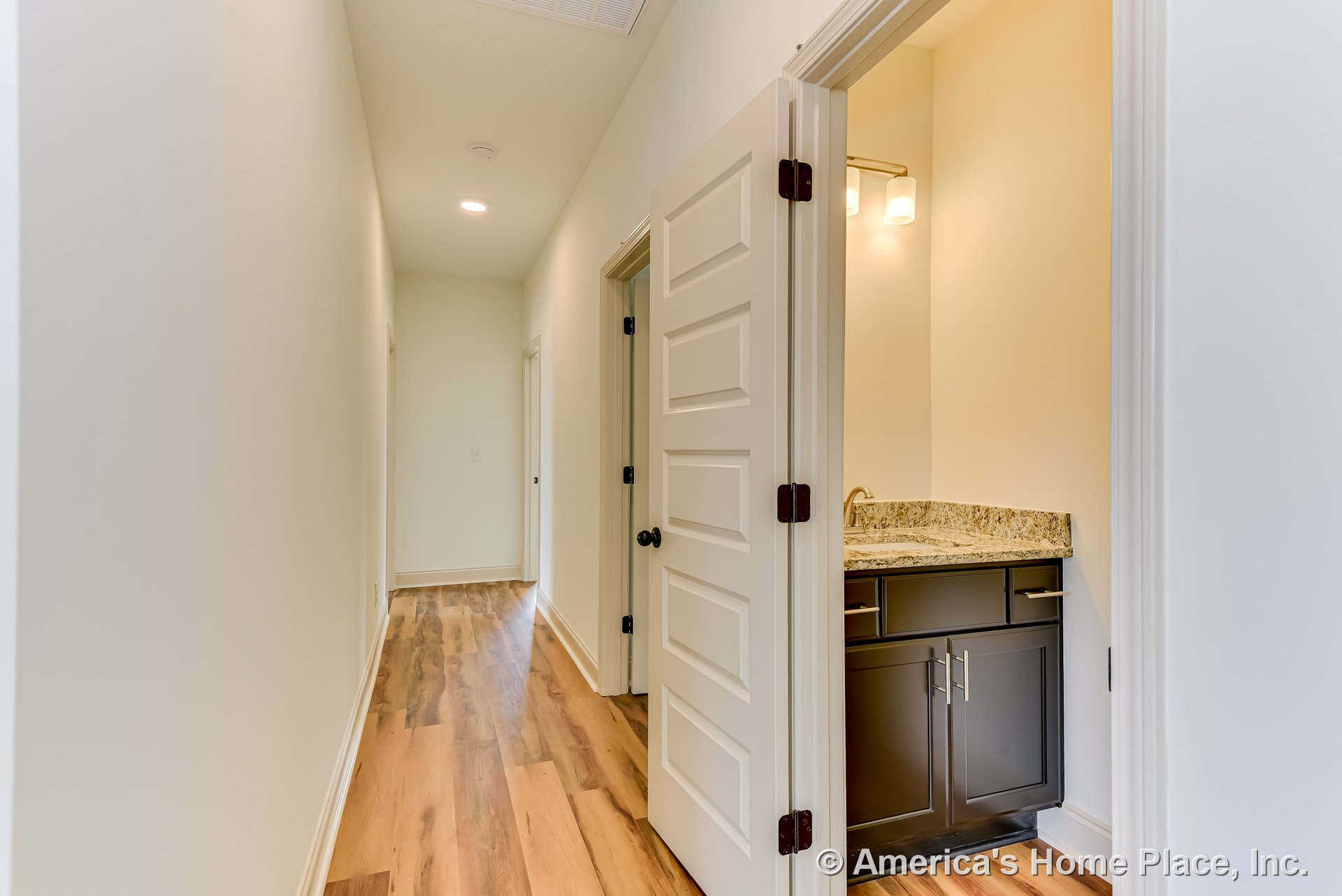 Hallway with recessed lighting, light wood plank flooring, and white paneled interior doors leading to a bathroom with granite countertop, dark vanity cabinetry, brushed nickel
