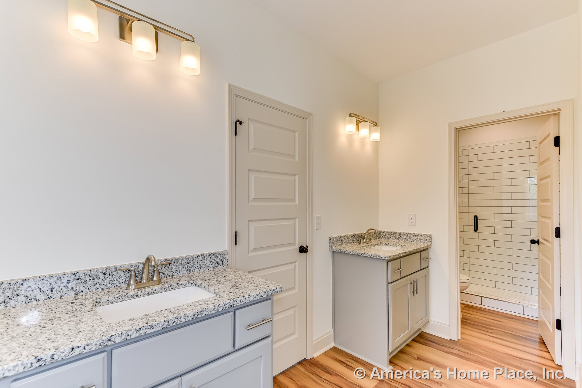 Dual vanities with granite countertops and white shaker cabinets, walk-in shower with white subway tile, wood-look flooring, and modern wall-mounted lighting fixtures.
