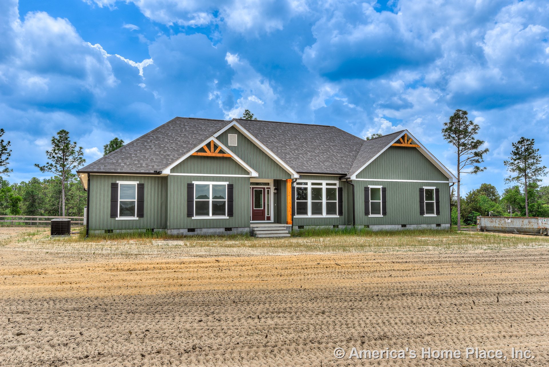 Green board-and-batten siding with white trim, decorative wood gable brackets, covered front porch, multiple double-hung windows, dark roof shingles, entry steps, and rural