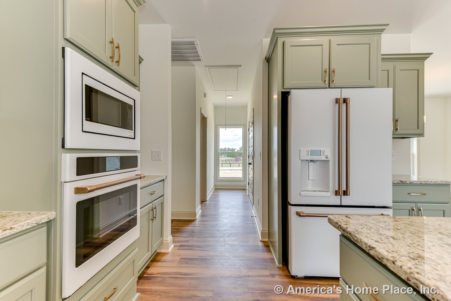 Sage green cabinets with granite countertops, built-in microwave and oven, white refrigerator with water dispenser, wood-look flooring, large window at hallway end.
