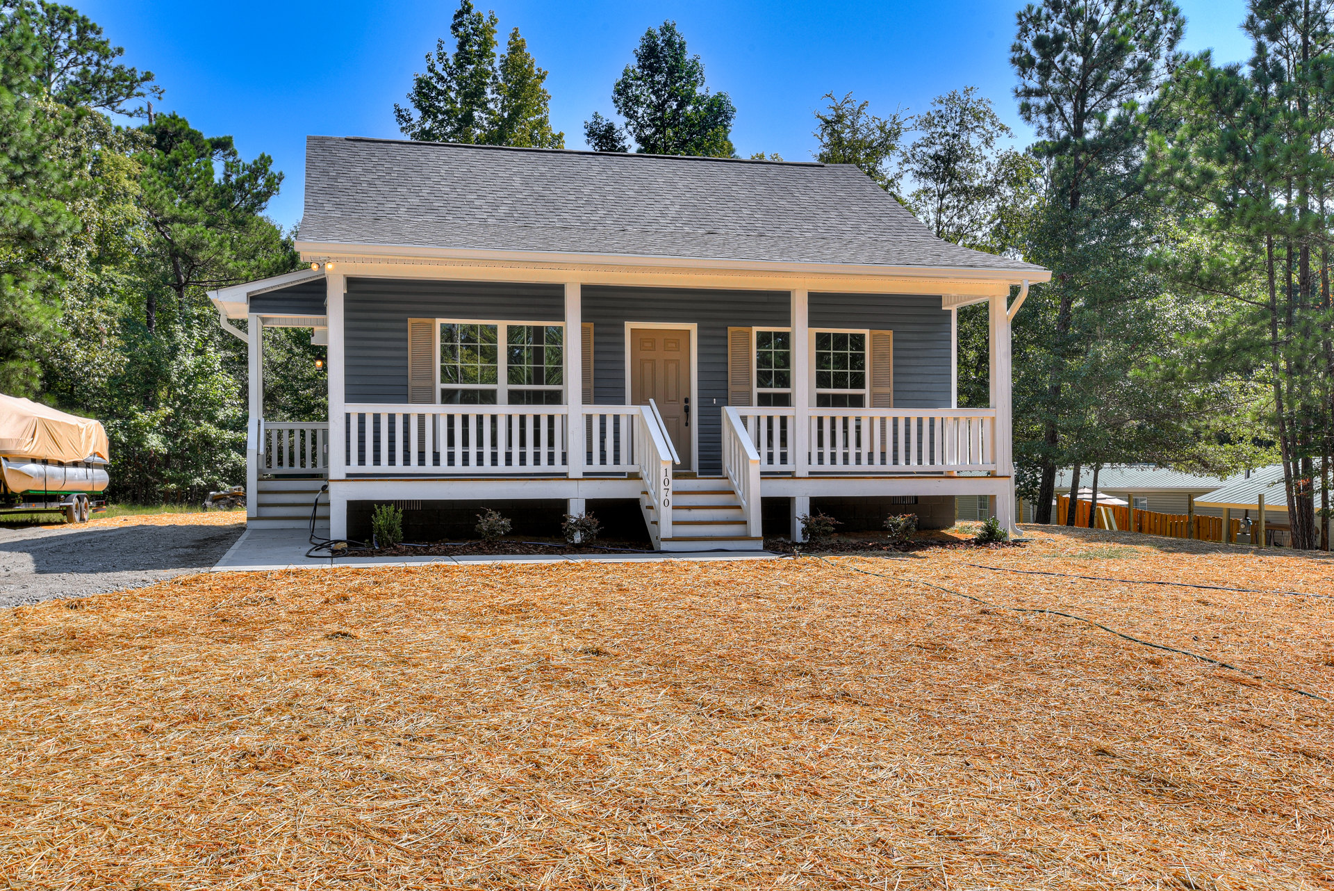 Two-story home with white wooden porch, brown front door, white railings, stairs leading to yard, pile of straw near steps, leafy tree and trailer with tarp in front yard