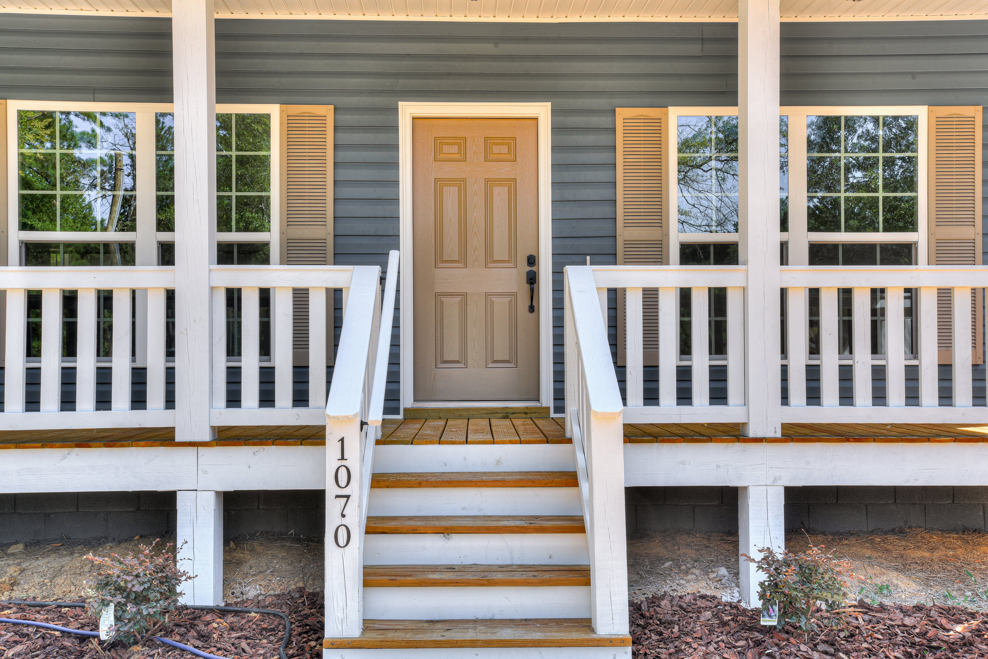 Brown front door with glass panels, white porch railings and stairs, white pillars, large window reflecting trees, wood plank siding