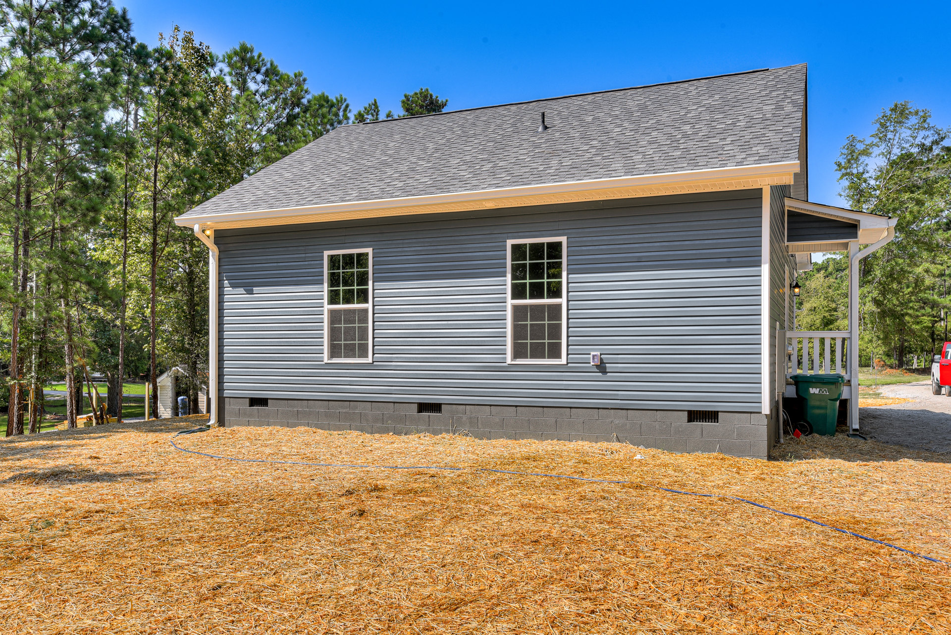 Blue metal roof with chimney, white-framed window, wood chip mulch covering yard, green wheeled trash can, blue garden hose on grass, mature trees surrounding cottage-style home
