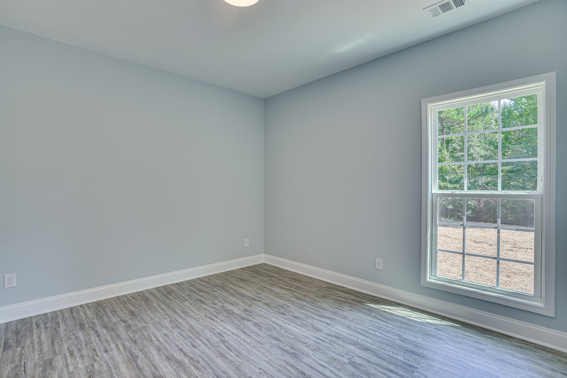 Sunlit room with multi-pane window, white plaster walls, wood flooring, ceiling light fixture, and view of hay field outside