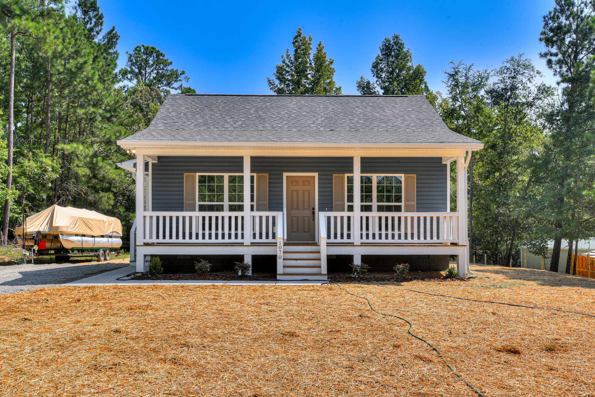 Two-story home with covered front porch, white railings, white door, large windows, manicured lawn, mature trees, and a boat on a trailer in the driveway