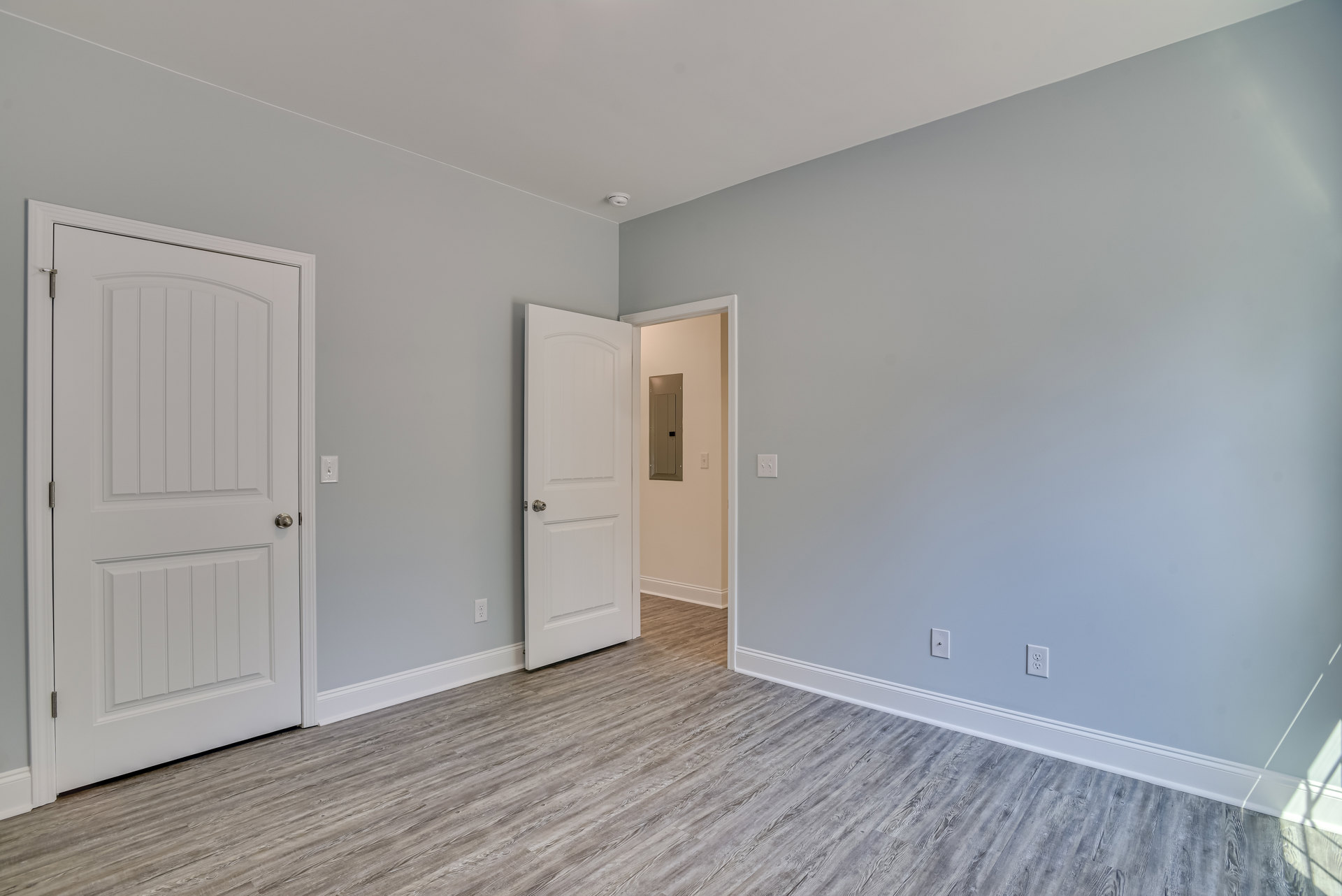White-painted room with two open doors, silver door knobs, and light wood laminate flooring; rectangular thermostat mounted on wall.