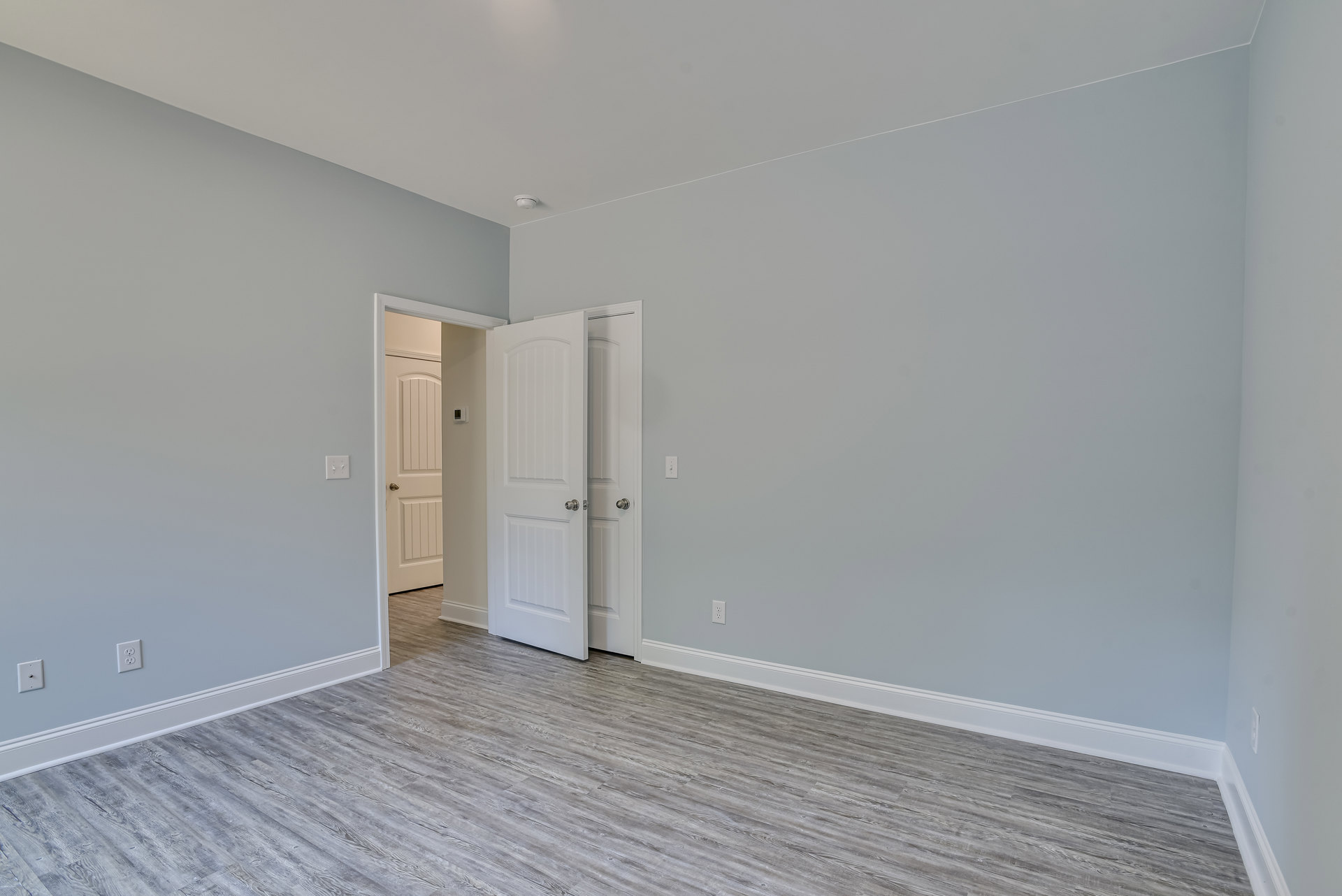 White-painted door open onto wood floor, revealing second open door; white ceiling with recessed light, neutral walls.