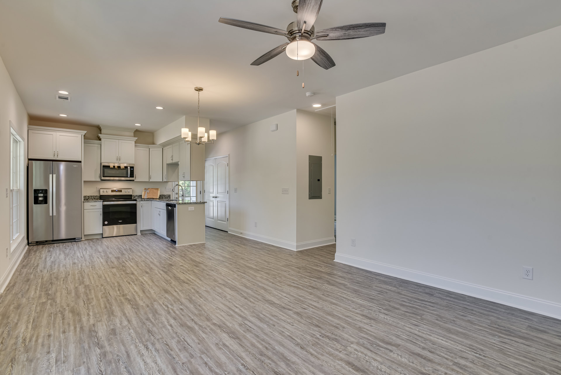 Open living room with wood flooring, white walls, ceiling fan with light fixture, adjacent kitchen featuring stainless steel refrigerator, stove, oven, and microwave