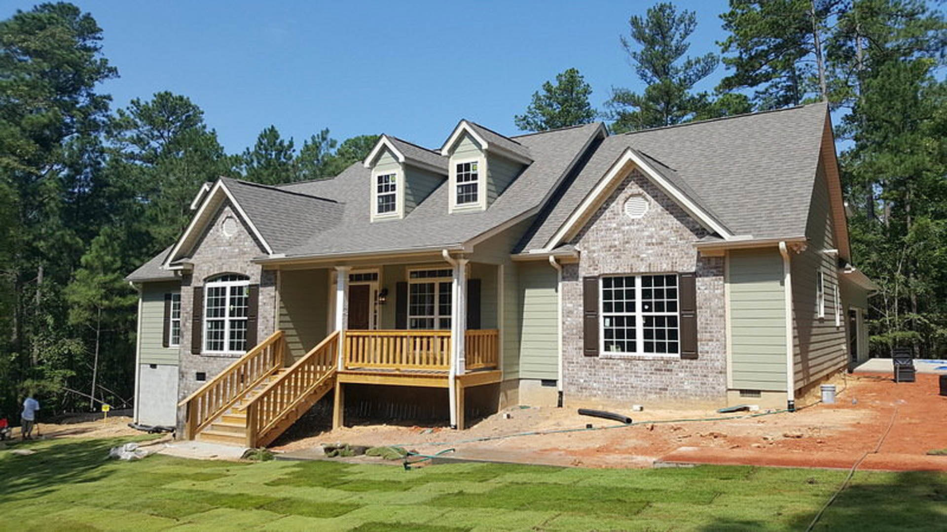 Two-story house with gray siding, white trim, covered wooden porch supported by white posts, manicured green lawn, large windows, and mature trees in the background