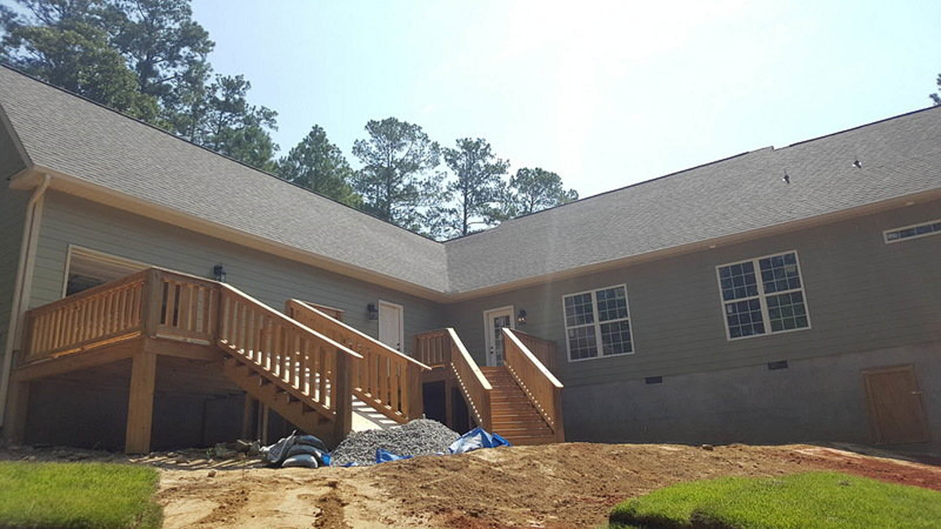 Two-story home with light siding, large square windows, wooden porch stairs, and a pile of dirt near the entry; trees and sky in the background.