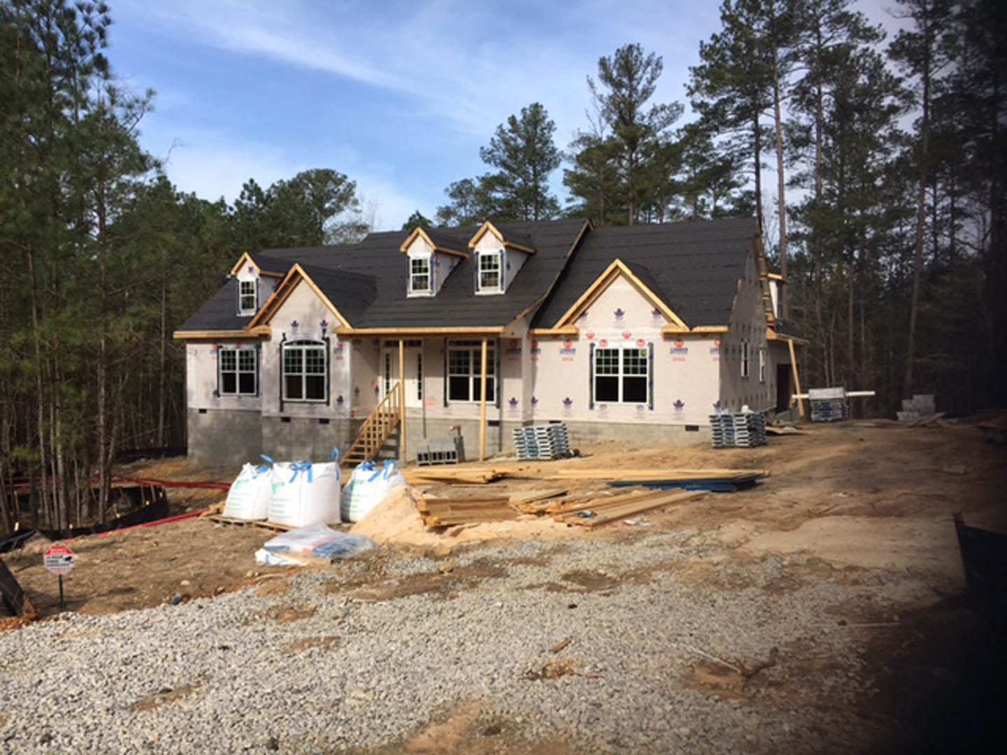Partially built house with black roof surrounded by tall trees, construction materials and blue ribbon bags scattered on dirt ground, cloudy sky overhead