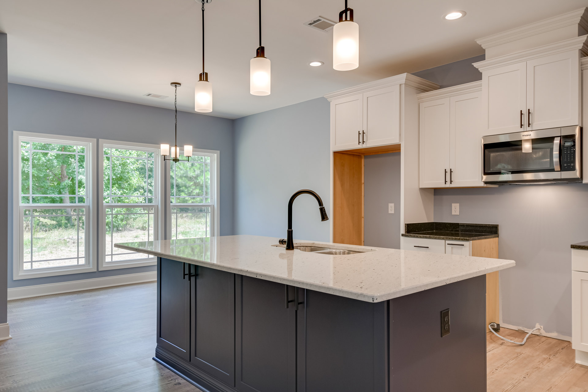 Modern kitchen featuring a central island with built-in sink and chrome tap, white cabinetry, stone countertops, stainless steel microwave with overhead lighting, and recessed