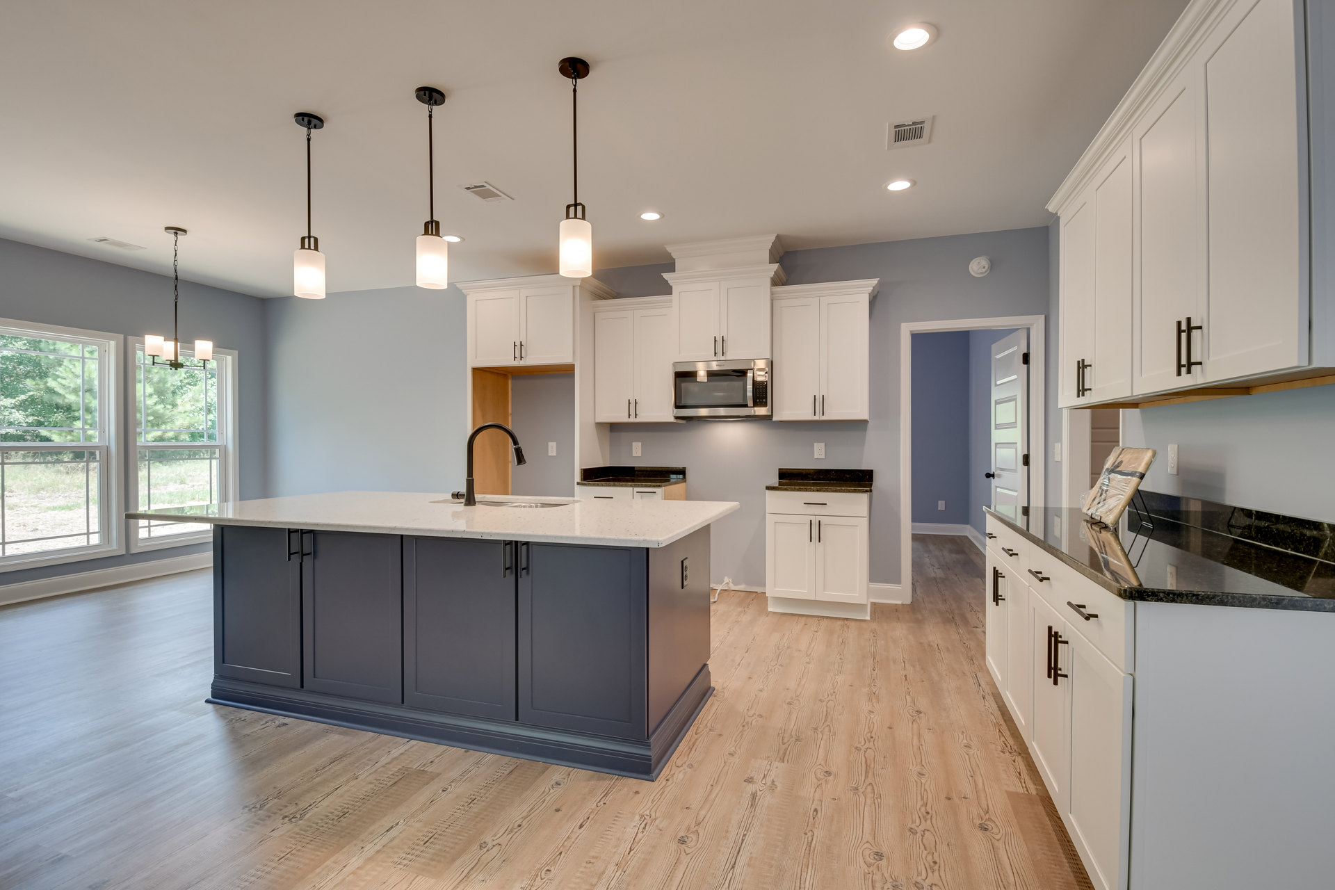 Kitchen with wood flooring, central island featuring a sink, white cabinetry with black countertops, built-in microwave, pendant lighting, and window overlooking trees.