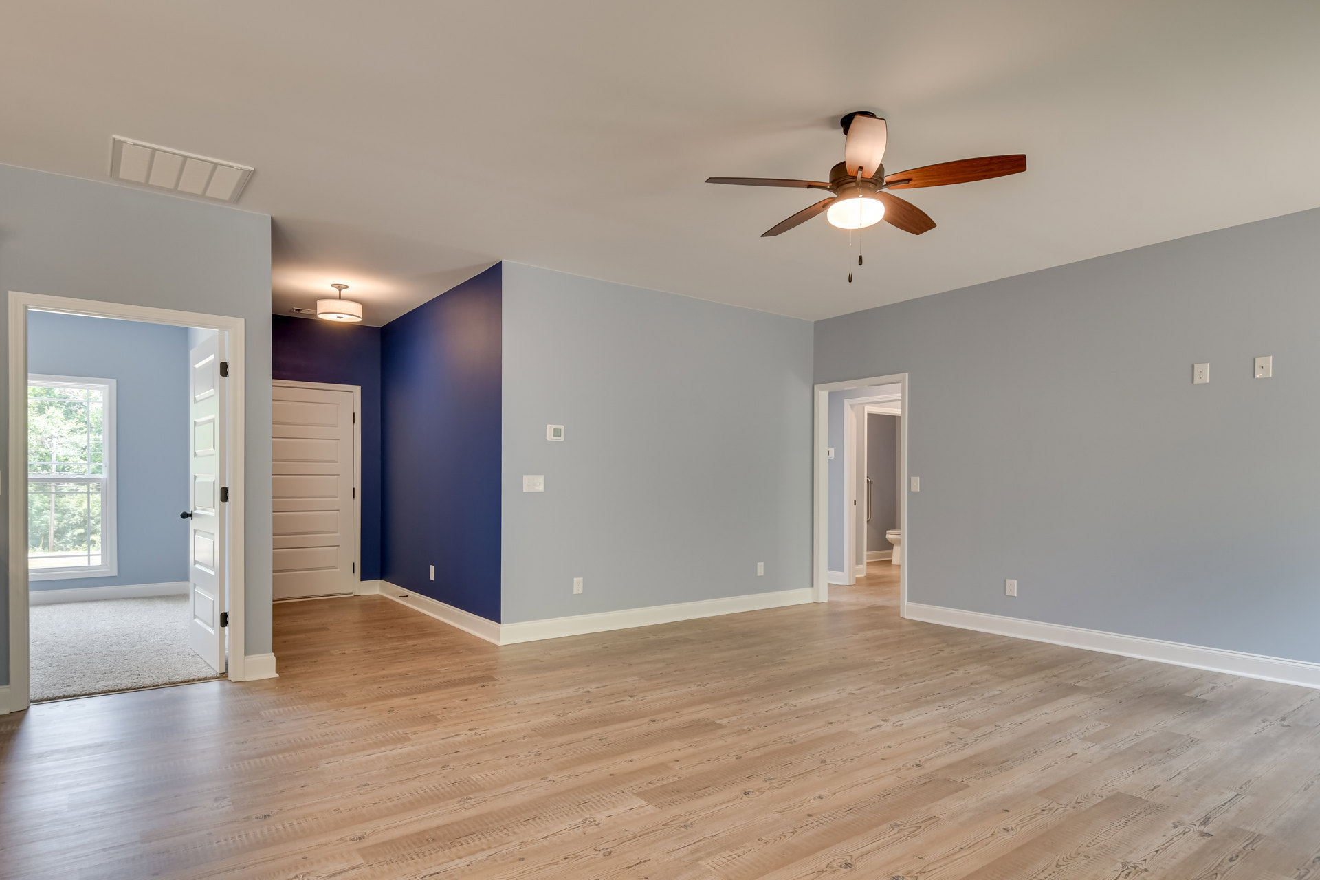 Ceiling fan with light fixture, wood laminate flooring with white trim, blue accent wall, white door, window overlooking trees, white grid-patterned ceiling