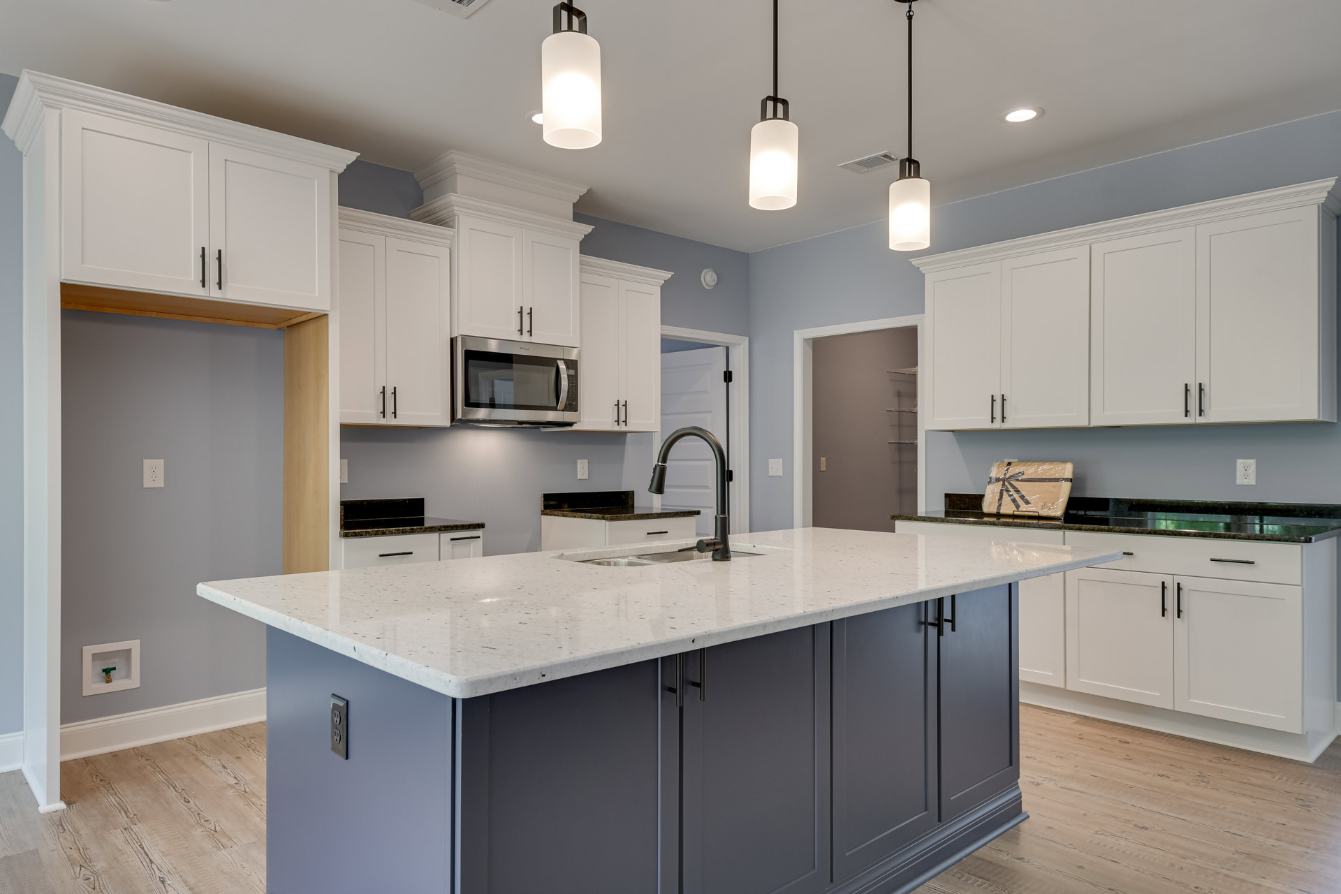 Spacious kitchen featuring a large central island with built-in sink, open microwave oven, white cabinetry, stone countertops, illuminated pendant light, hardwood flooring, and