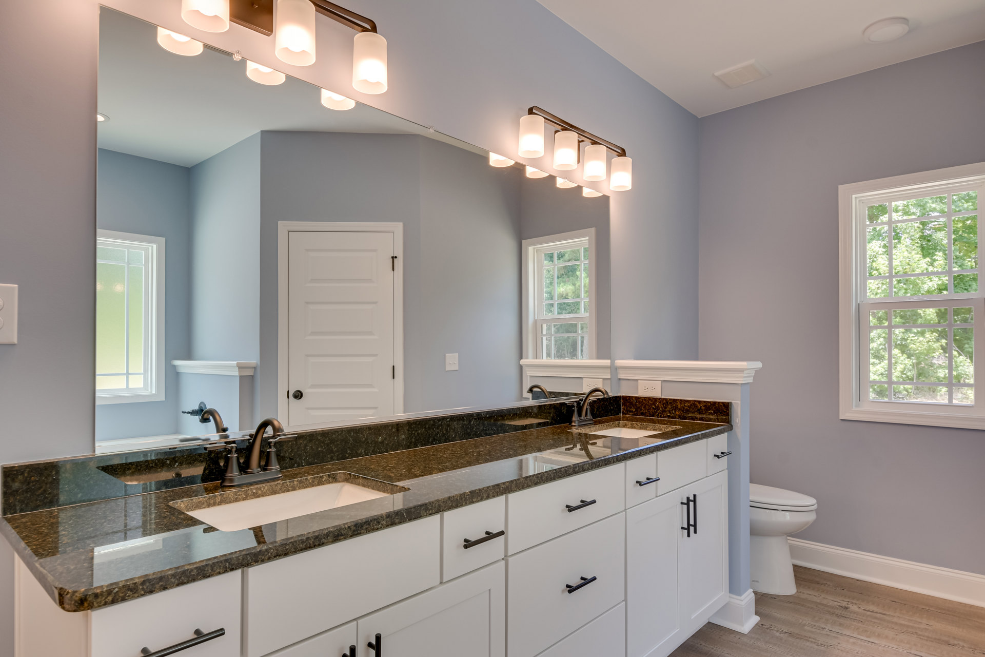 Modern bathroom featuring a rectangular mirror above a white sink with chrome faucet, light-colored tile backsplash, wood cabinetry, white toilet, and a window overlooking green
