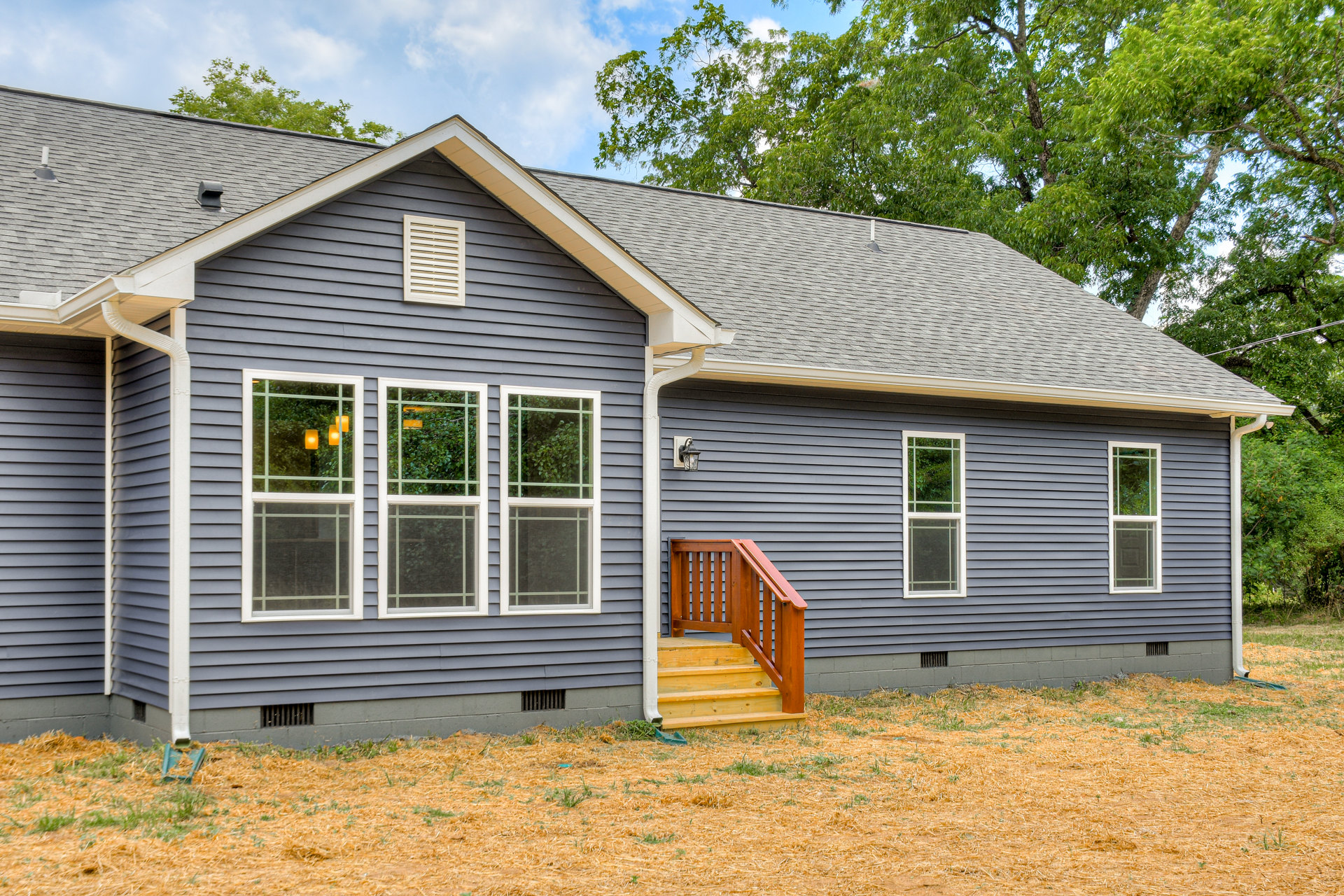 Front porch with white railings, wooden staircase leading to entrance, white-framed windows, green bushes and trees surrounding exterior, cloudy sky overhead