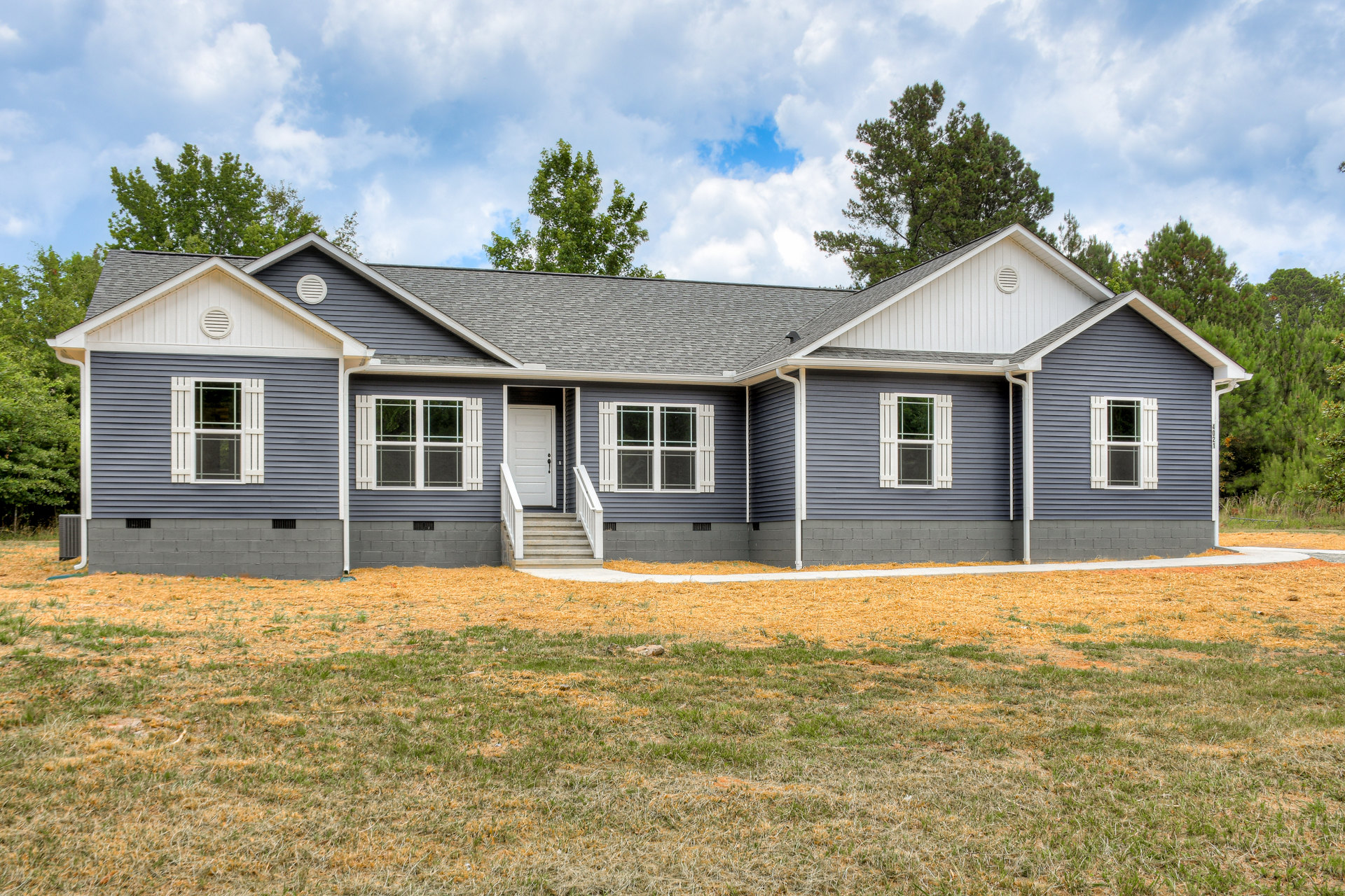 Two-story house with white door, white-trimmed windows, brick foundation, front staircase, and green lawn bordered by mature trees