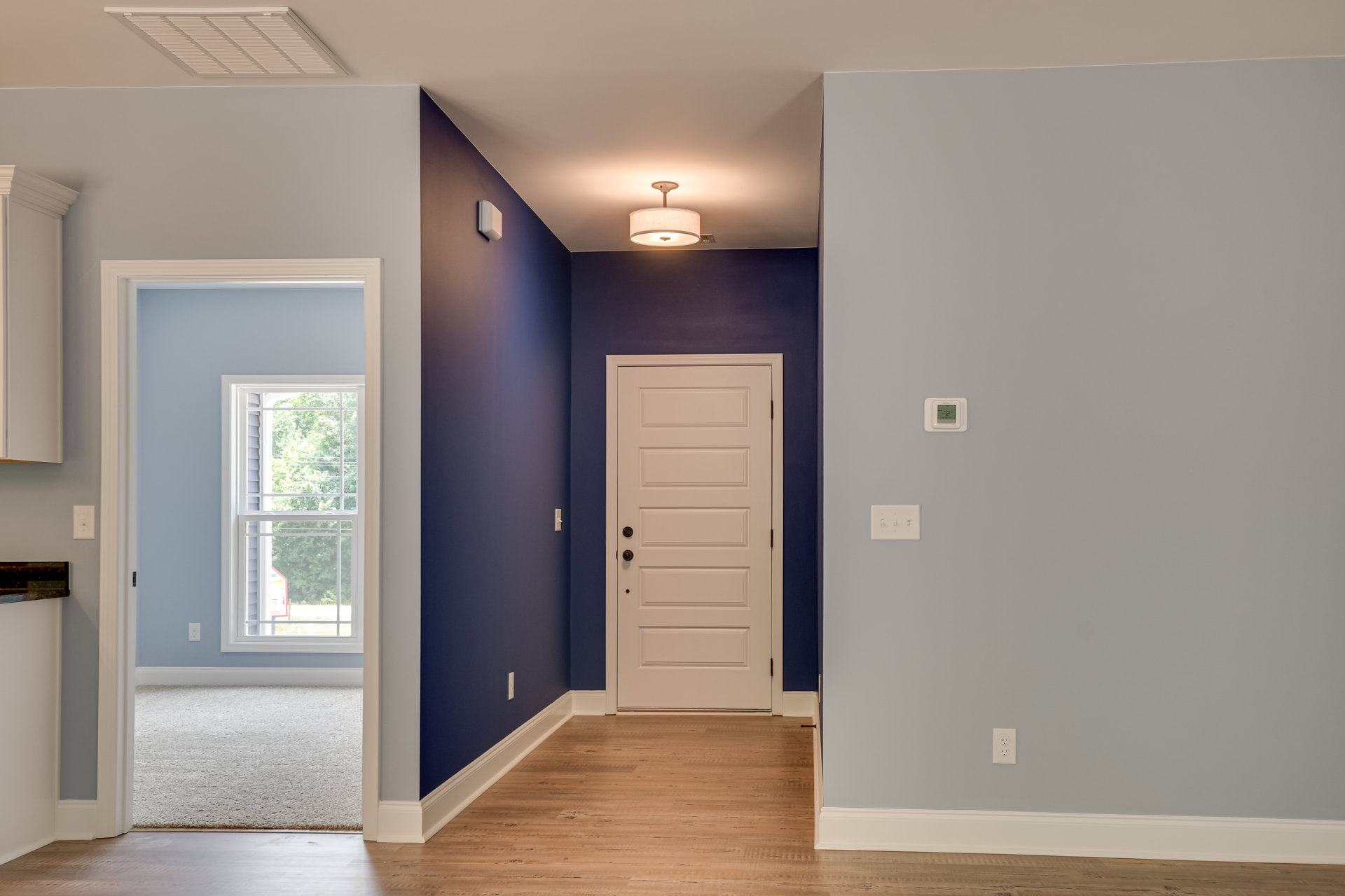 Hallway with blue walls, white door featuring black knobs, window overlooking trees, white ceiling with vent, white wall with light switch, light fixture with white shade