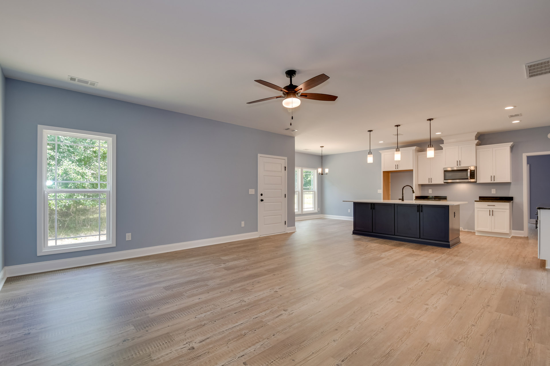 Open kitchen with white island, wood flooring, ceiling fan with light, large window showing trees, and white door with black hardware