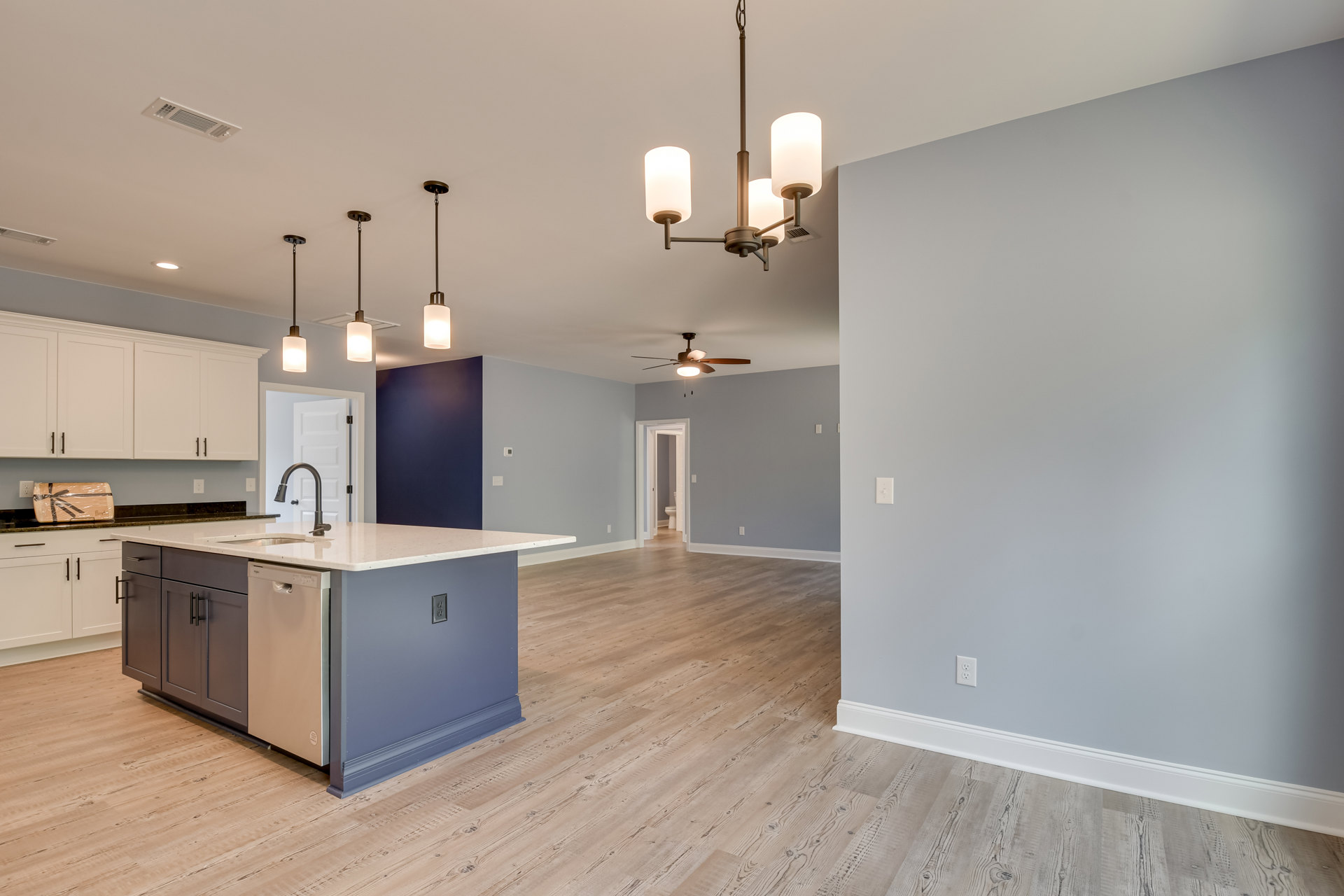 Open kitchen and dining area with wood flooring, ceiling fan, kitchen island featuring a sink, white cabinetry, wall-mounted light, light switches, white door with matching frame