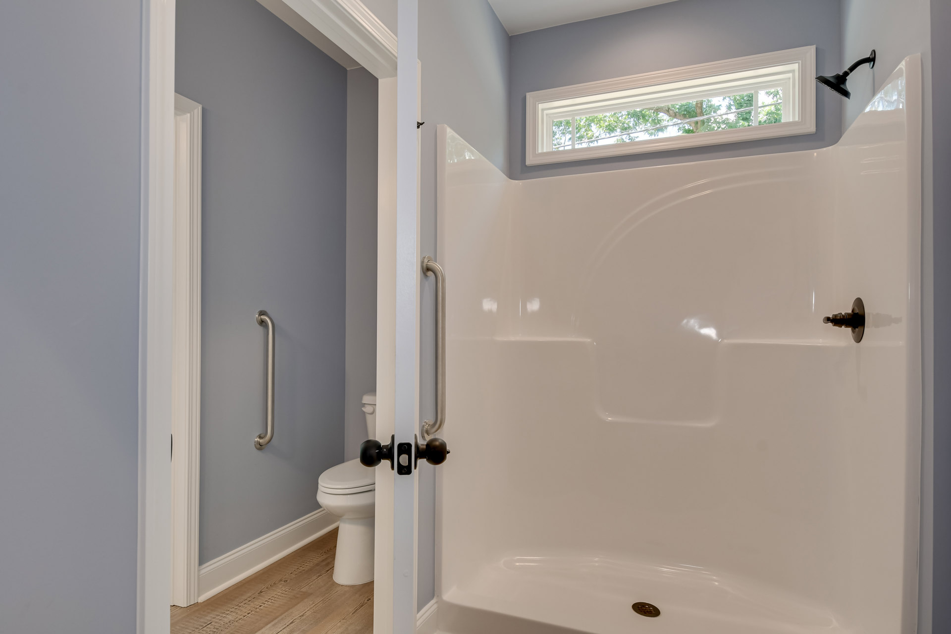 Modern bathroom featuring a glass-enclosed shower with white tile walls, a white toilet with closed lid, black wall-mounted lamp, chrome door handle, and a window overlooking leafy