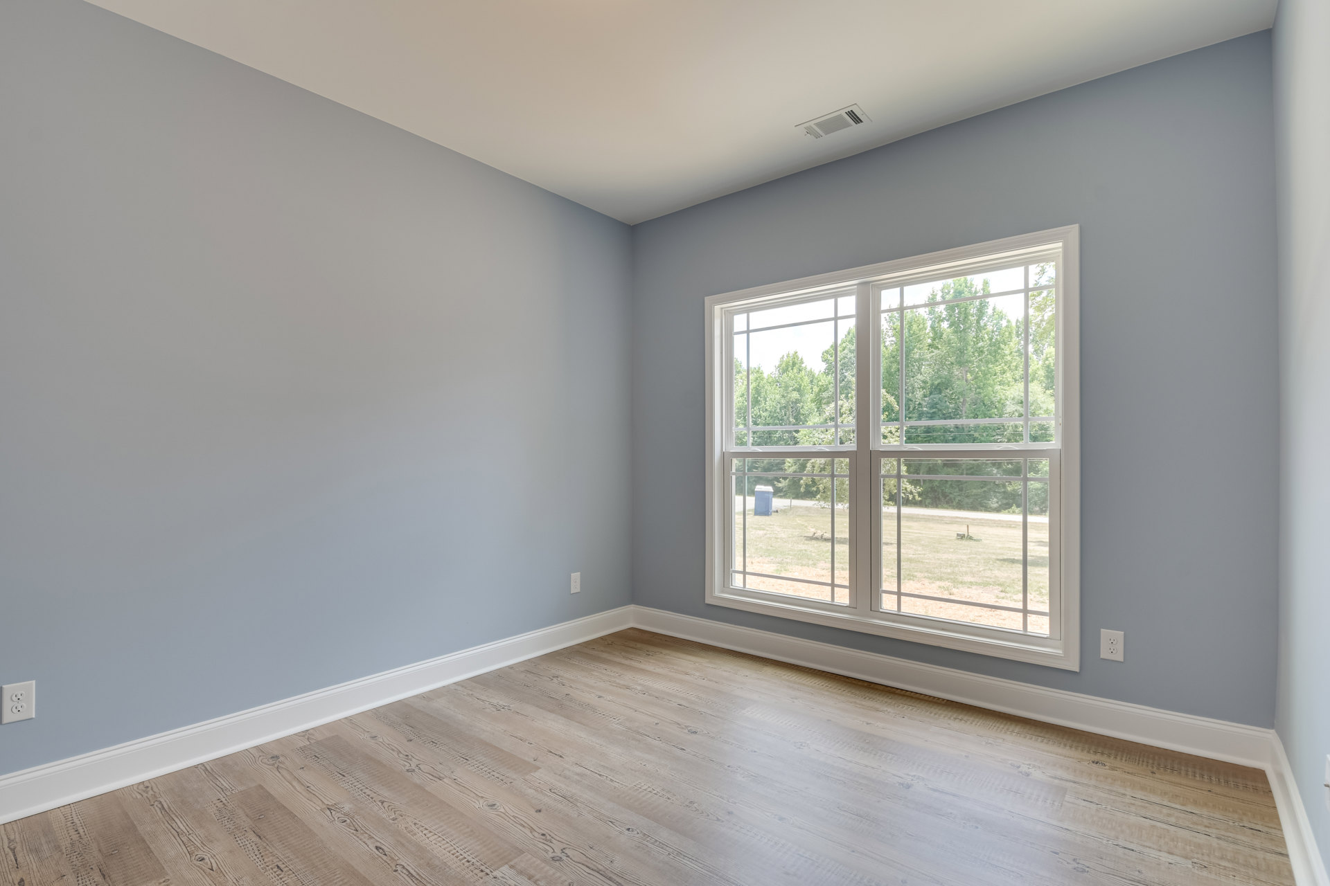 Sunlit room featuring large window with view of trees, smooth hardwood flooring, white plaster walls, ceiling vent, and wooden door.