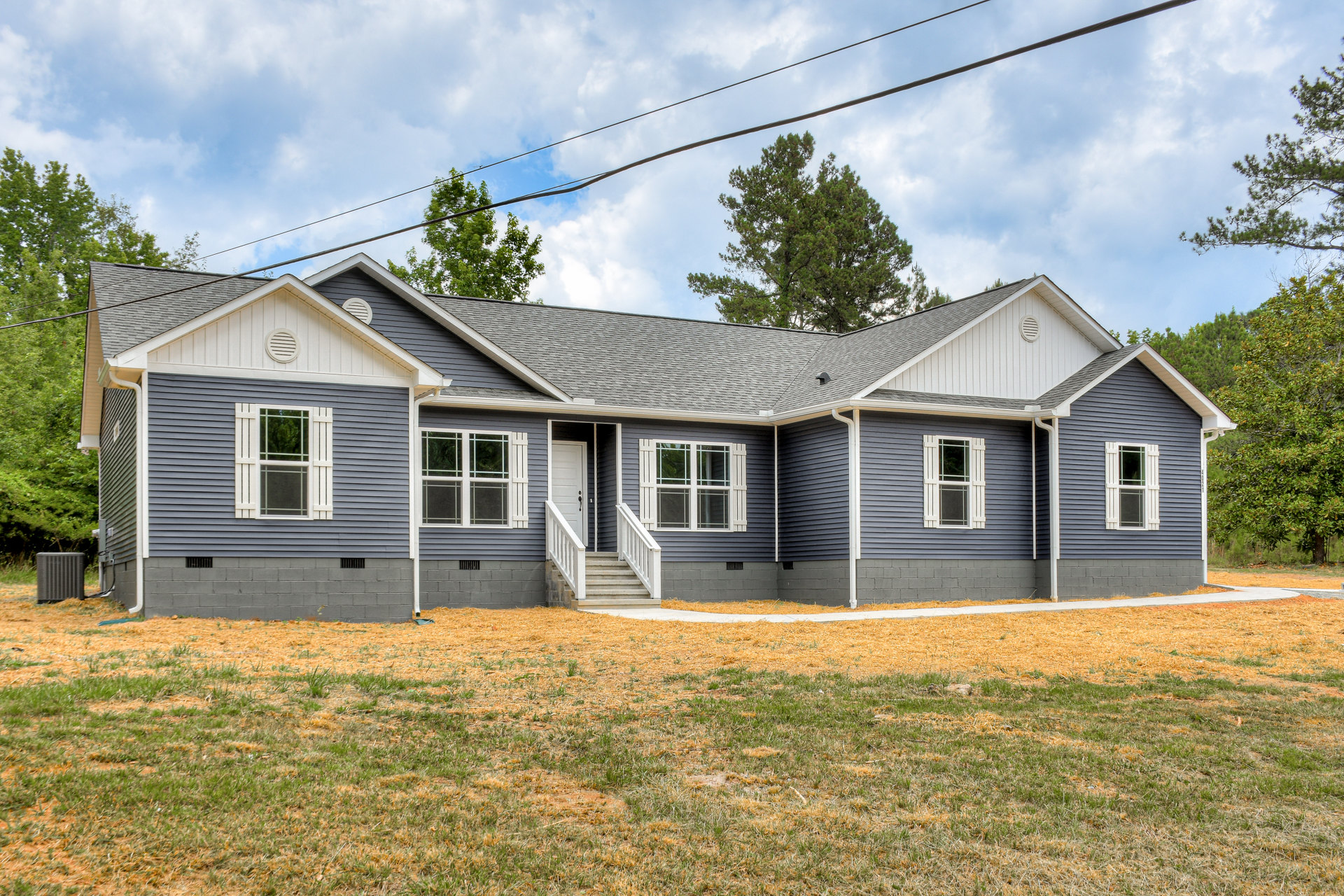 Two-story home with white door and stairs, white-trimmed windows, grey utility box on green lawn, power lines overhead, mature trees in background, cloudy sky